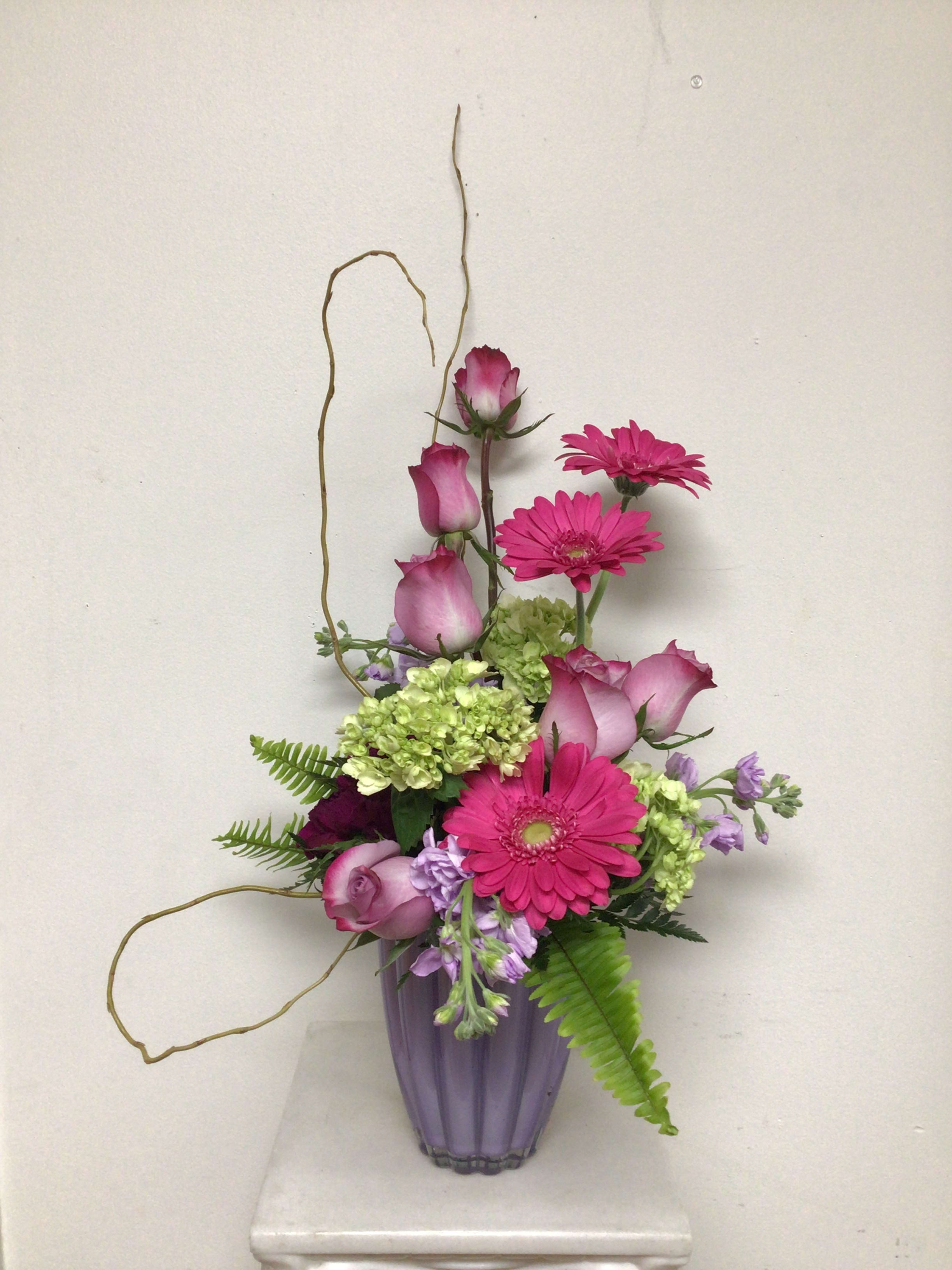 Trio - This contemporary arrangement is arranged in a lavender clam shell vase and  is filled with dark purple carnations, lavender roses, green hydrangea, lavender stock, pink gerberas, sword fern, mixed greenery and accented with curly willow, all done in groupings of three. Approximate size is 22 inches tall and 15 inches wide.