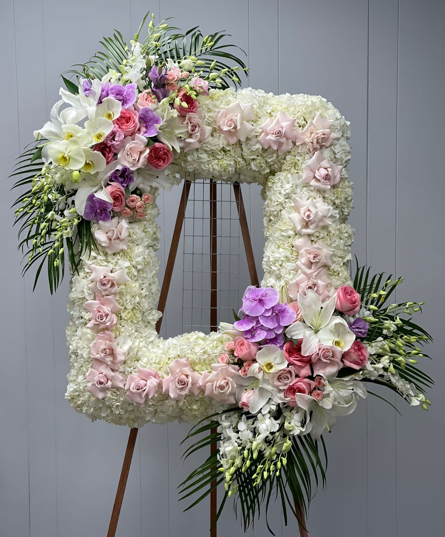 Memorial Frame Spray 1 - This luxurious frame spray is designed by Kenneth Village Flowers as an addition to any memorial service. This arrangement features majestic white hydrangeas that will ease the minds and comfort the souls that are grieving with lilies, orchids and roses to represent the vibrance of their life. 