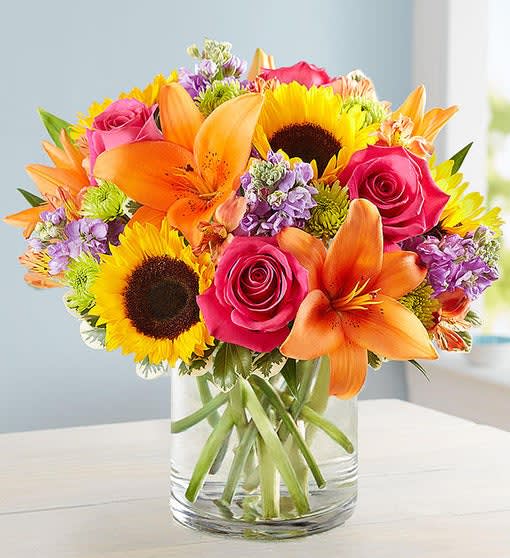 FLORAL EMBRACE FLORAL EMBRACE - All-around arrangement of hot pink roses and carnations; orange Asiatic lilies and Peruvian lilies (alstroemeria); yellow sunflowers; lavender stock; purple monte casino; green Athos poms; accented with assorted greenery