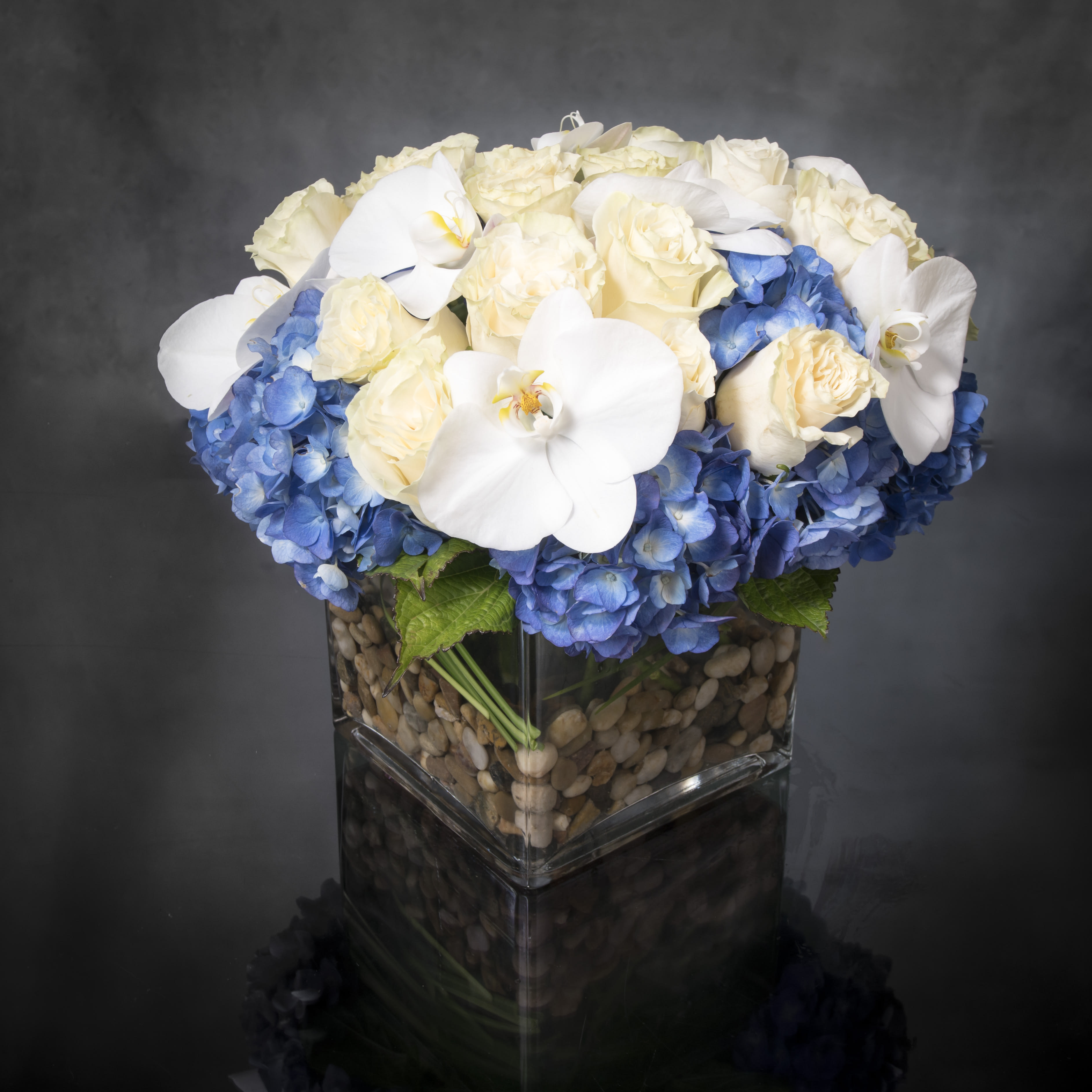 Heart of the Sea  - This beach-inspired bouquet captures the blissful beauty of a seaside retreat. Bunches of blooms in cool, soothing shades are hand-arranged in a glass cube vase. Everyone's favorite summer flowers are here - light blue Hydrangeas, white Phalaenopsis Orchids, and beautiful white Roses. It’s a gift that celebrates the carefree spirit of summer, while surprising the people you care about most.  Standard: 12 stems of roses  Deluxe: 24 stems of roses ( As Shown)  Premium: 36 stems of roses