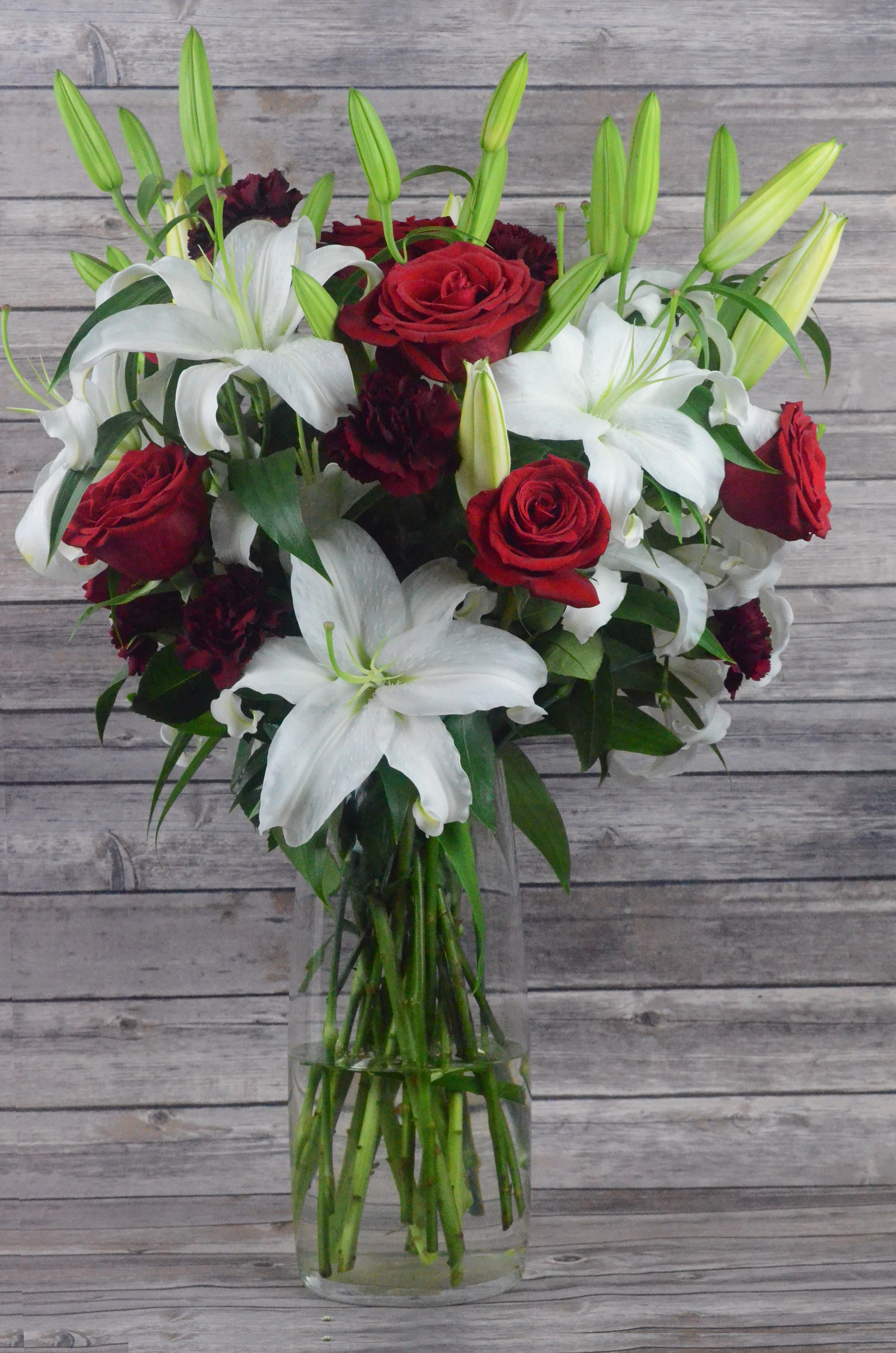 Majestic Love - This assortment of white lilies, red roses and red carnations are the way to get your point across in a big way.  All bouquets are arranged by our professional floral design team accented with seasonal filler (i.e. greenery, baby’s breath, etc.) inside an elegant glass vase or gift box. Blooms are hand delivered in person to enhance any celebration or a “just because” surprise. When artistically designed in a clear glass vase arrangement measures approximately 23&quot;H x 18&quot;L.