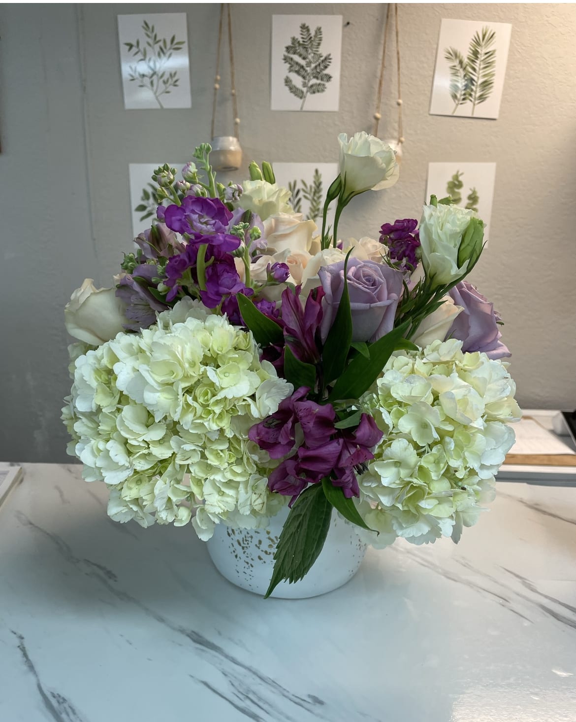 Elegant but mighty - Our unique modern white with gold speckled vase with a mix of purple and pink blooms Delicately placed on a bed of white hydrangea.