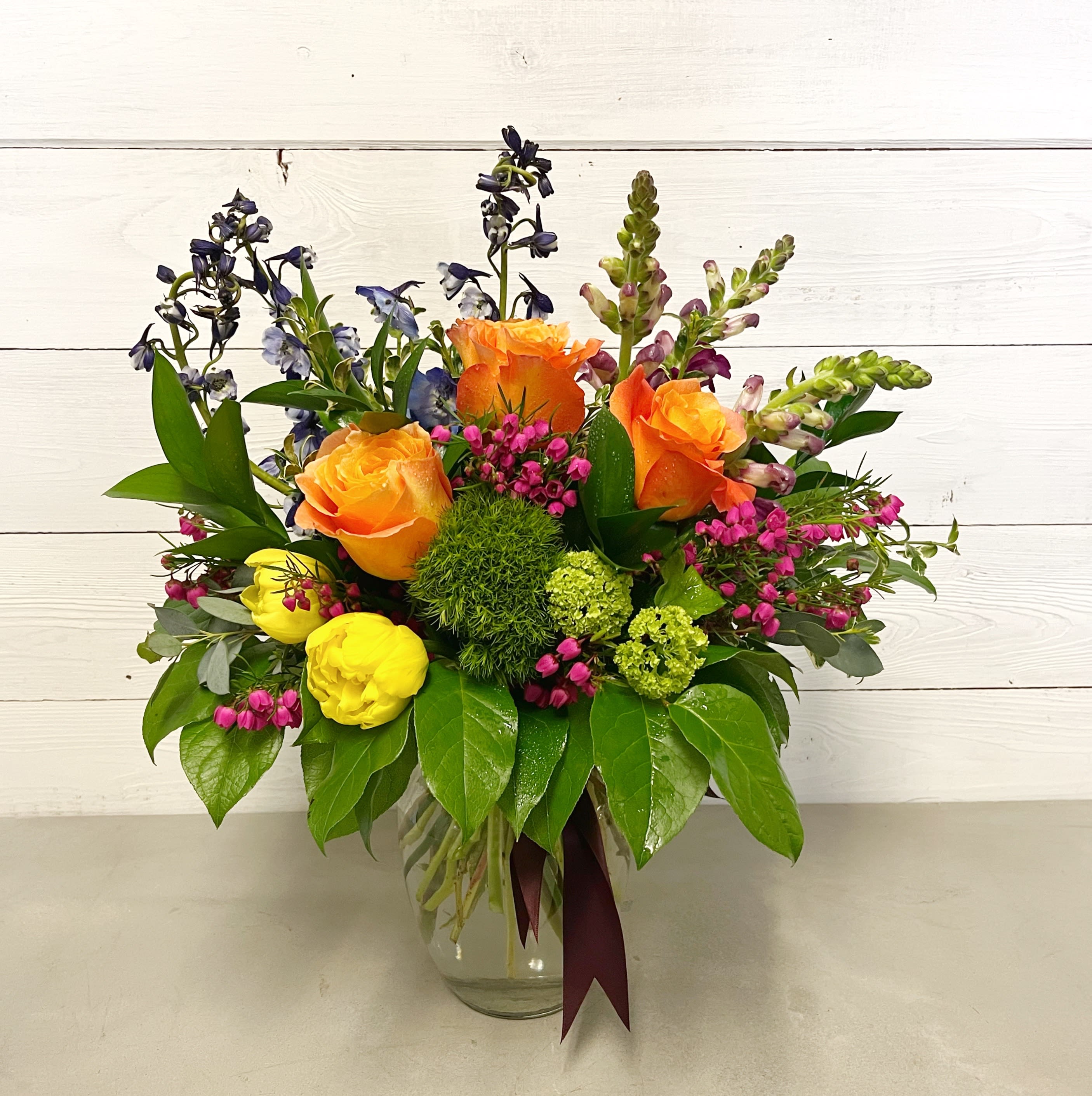 May - This arrangement features a bright mix of roses, snapdragons, delphinium, tulips, dianthus, greens, and filler in a clear glass vase. Please be advised, that the flowers and vase may vary. 