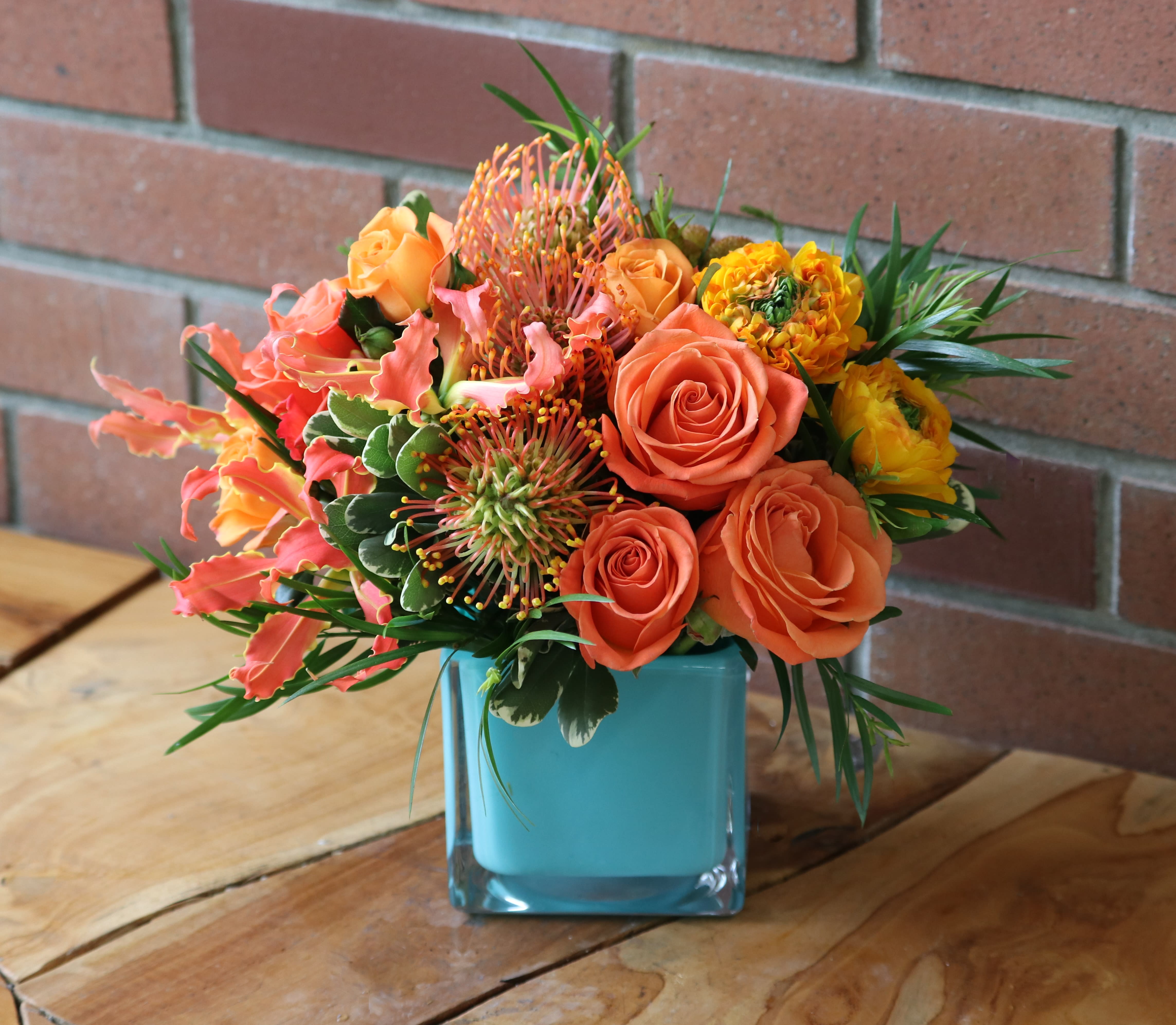 Color Spark Bouquet - Beautiful blue cube vase with high end orange and yellow blossoms. tropical pin cushion proteas, orange roses, bi color yellow ranunculus, gloriosa, premium floral greenery arranged tight in a low glass cube vase.  Deluxe &amp; Premium get more of the same flowers in a larger sized blue cube vase.