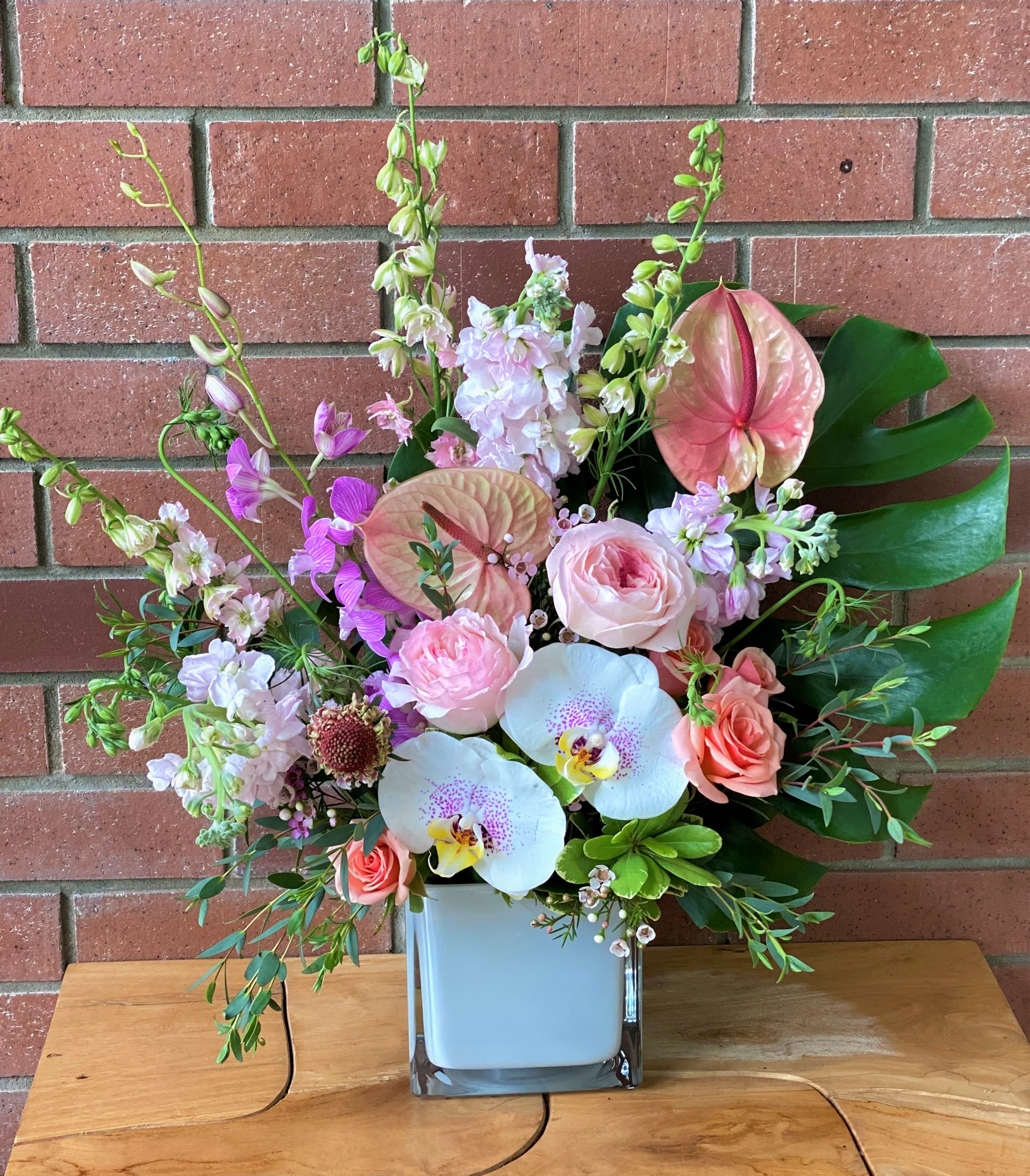 Tropical Fantasy  - Gorgeous arrangement full of premium blooms: Orchids, dendrobium orchids, anthurium, larkspur, scented stock, scented garden roses, spray roses, monstera leaves arranged with premium greens and fillers in a square 5x5x5 white glass vase.