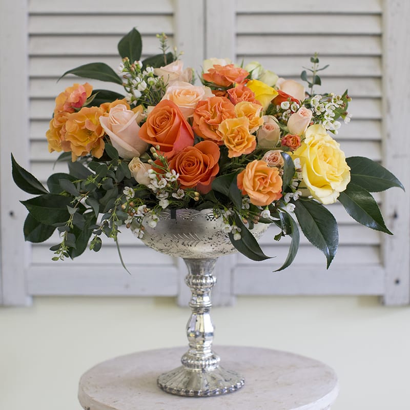 Rose Elegance - A mix of orange, peach and yellow roses and spray roses accented with seeded eucalyptus and wax flowers in a tall mercury glass compote. 