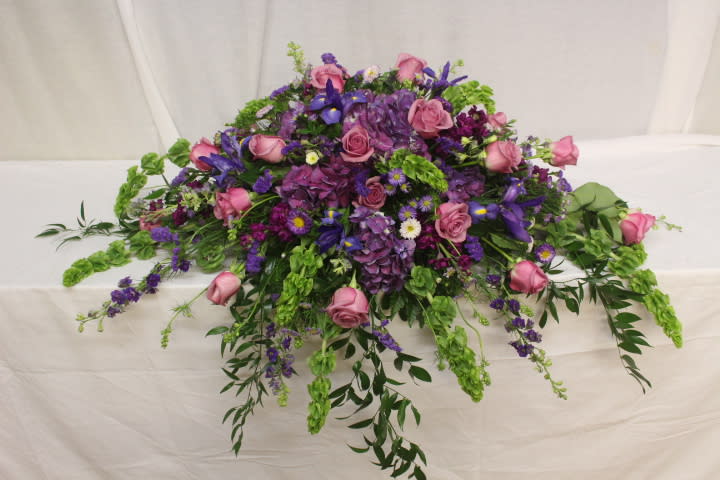 TB-2122 Peaceful Tribute Casket Spray - The Peaceful Tribute Casket Spray is an exquisite presentation to honor the life of the deceased. The color palette of blue, lavender and purple hues include iris, roses, hydrangea,stock, larkspur and asters, all of which are surrounded by fresh, beautiful bells of ireland and soft tendrils of italian ruskus. The culmination is a perfect display to express your devotion to your loved one.