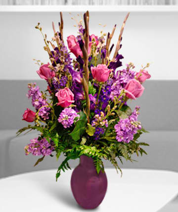 Purple Pride - Gladiolus, stock, &amp; larkspur in purples with accent of pink roses.  Substitutions of equal or greater value may be made depending on season and availability of product or container. 