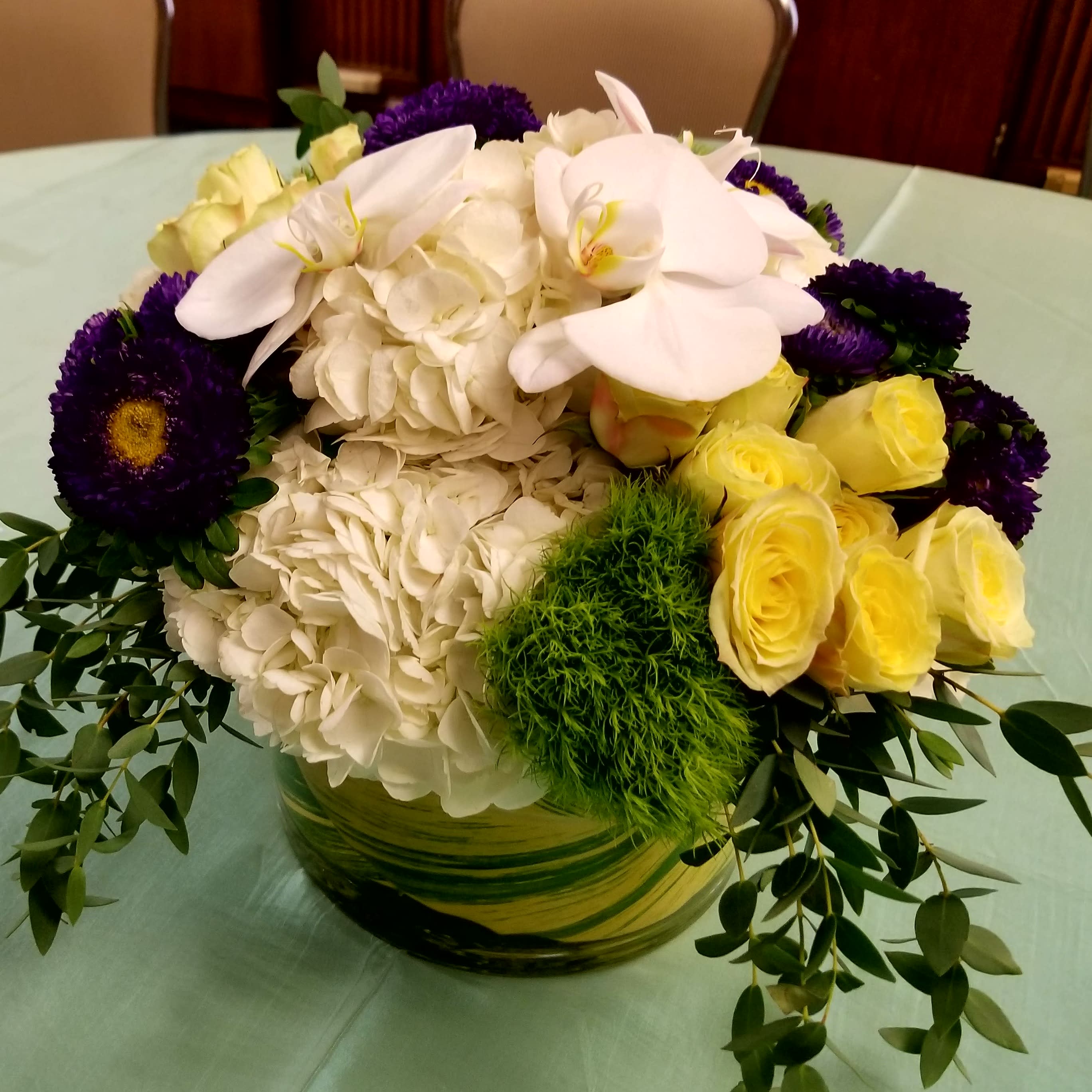 Braun Standard - Hydrangea, trick, spray roses &amp; phalaenopsis orchids in a leaf wrapped glass container.