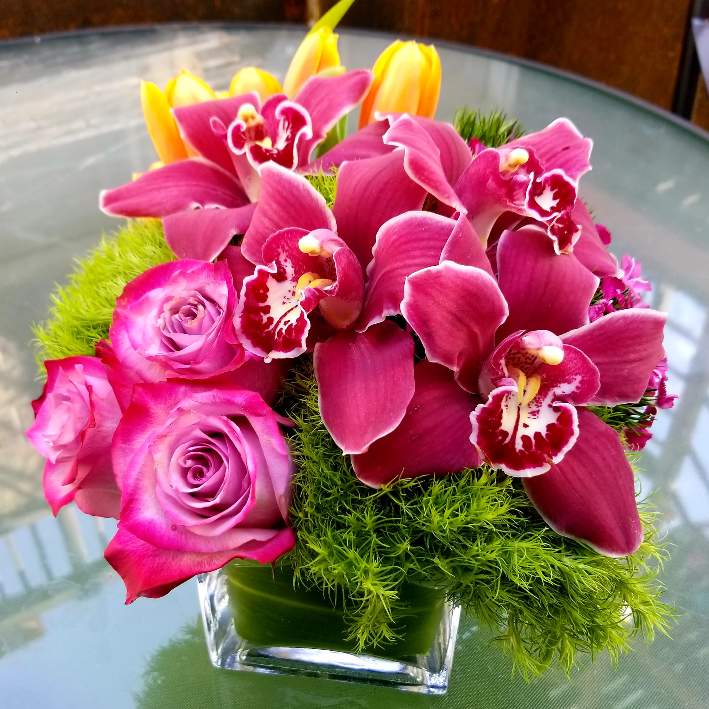 Raspberry Trick - Trick, roses, tulips &amp; cymbidium orchids in a glass leaf wrapped cube.