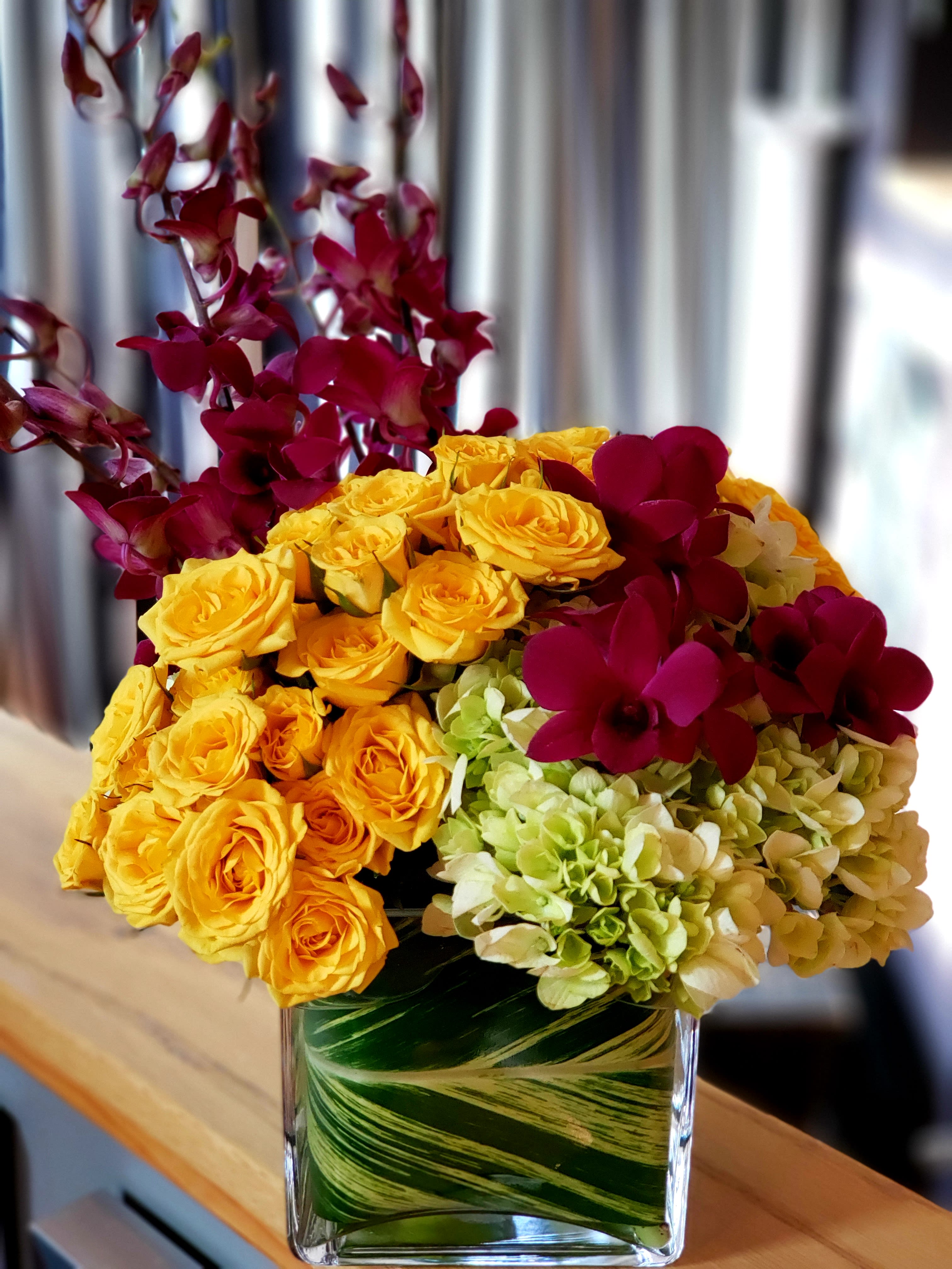 Vallarta Sunrise - Hydrangeas, spray roses &amp; dendrobium orchids in a ti leaf wrapped square glass container. 