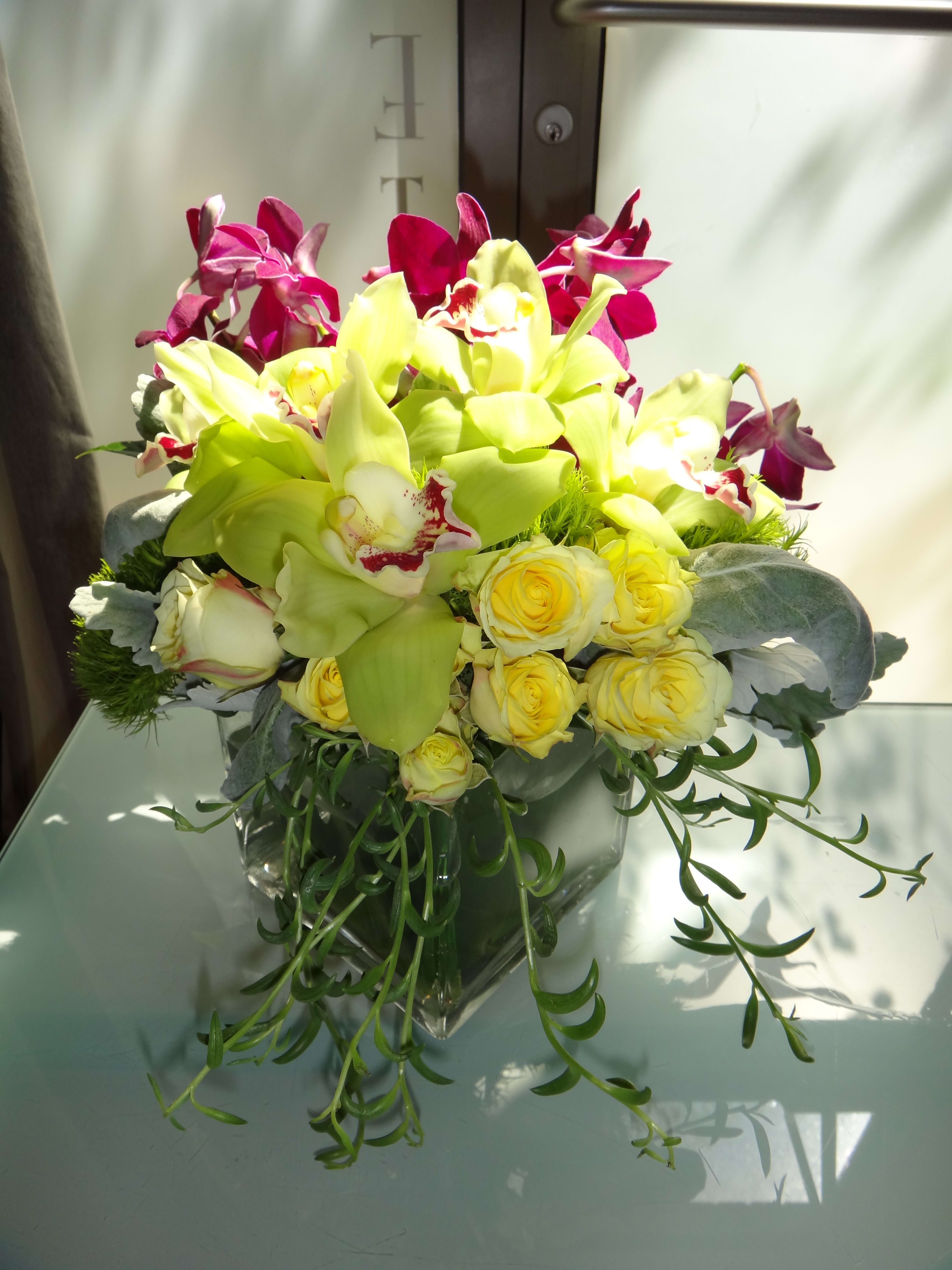 Sunrise - Spray roses, dusty miller, trick, &amp; cymbidium orchids in a square glass container.