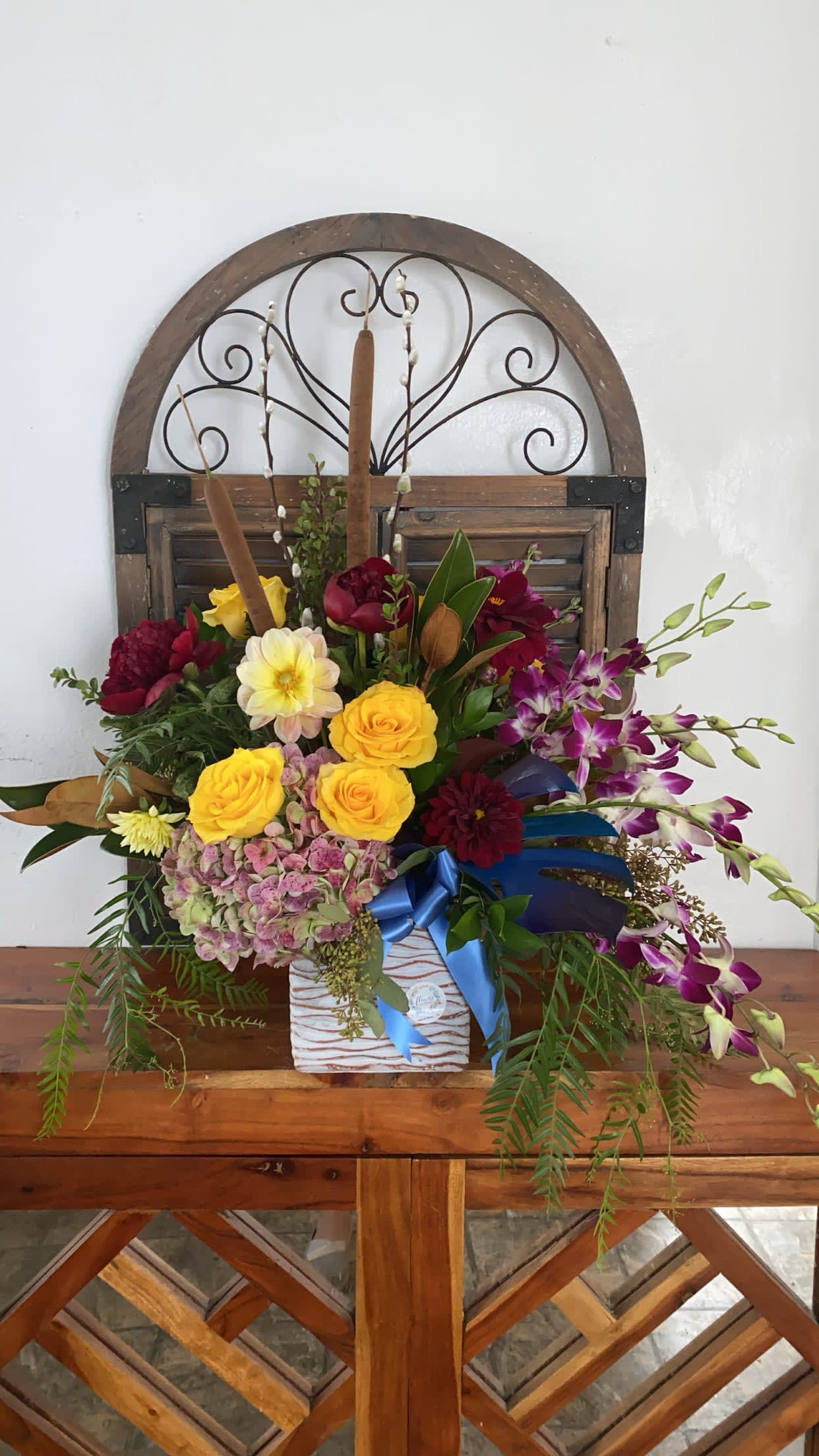 Love Trail - Amazing combination of deep burgundy peonies and dahlias, with yellow roses and orchids.Rustic cement vase and hangings tropical leaves.
