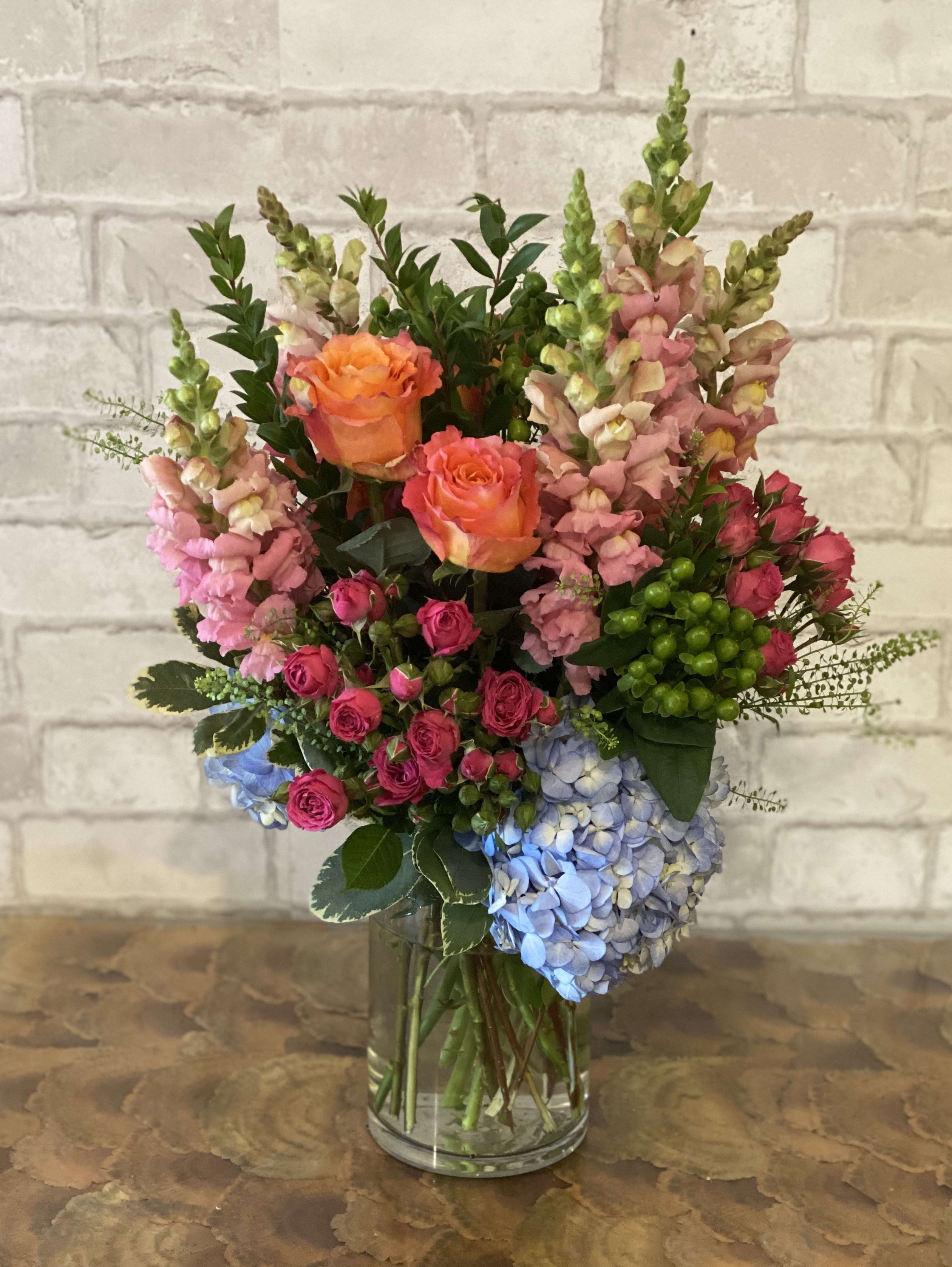 Spirited Snapdragon - An assortment of blue hydrangea, pink snapdragon, peach roses and hot pink spray roses. Arranged in a medium cylinder vase.