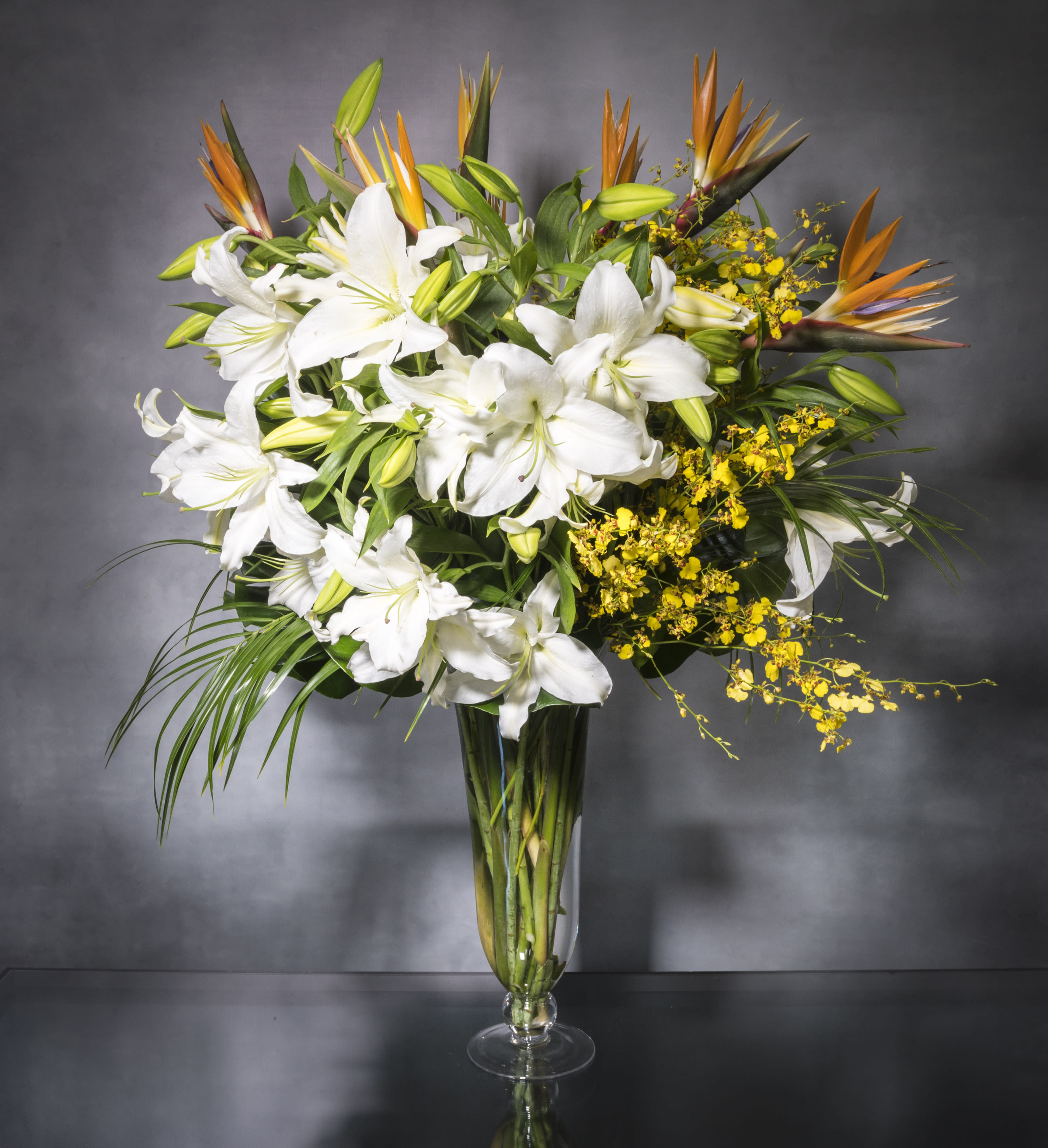 Tropical Vibrations - Imported White Lilies, bird of paradise and  dancing lady orchids which take center stage in this design. This delightful piece is a lovely choice for Mother's Day, Birthday, and all occasions.