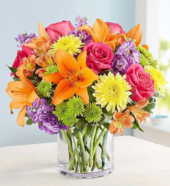 Floral Embrace™ - Like a warm embrace, our vibrant flower bouquet delivers your sentiments to someone special. A rich gathering of yellow and orange blooms, with pops of bright pink and purple, it's more than a gift - it's a way to express how you feel inside.  All-around arrangement of hot pink roses and carnations; orange Asiatic lilies and Peruvian lilies (alstroemeria); yellow sunflowers or similar blooms; lavender stock; purple monte casino; green Athos poms; accented with assorted greenery Artistically designed in a glass vase Large arrangement does not include carnations; measures approximately 13&quot;H x 13&quot;L; vase measures 6&quot;H Medium arrangement; measures approximately 12&quot;H x 12&quot;L; vase measures 5&quot;H Small arrangement does not include stock; measures approximately 11&quot;H x 11&quot;L; vase measures 5&quot; Our florists hand-design each arrangement, so colors, varieties and container may vary due to local availability To ensure lasting beauty, lilies may arrive in bud form, and will fully bloom over the next few days Sunflowers or similar blooms may arrive with light or dark centers Our local florists always put their passion and creativity into each gift. At times, they may need to make substitutions to floral arrangements Fruit Bouquets, or gift baskets to ensure your order is fresh and delivered in a timely manner. This is especially true as we navigate the evolving health crisis. Please know that in this instance, the utmost care and attention is given to ensure your gift is of equal value.