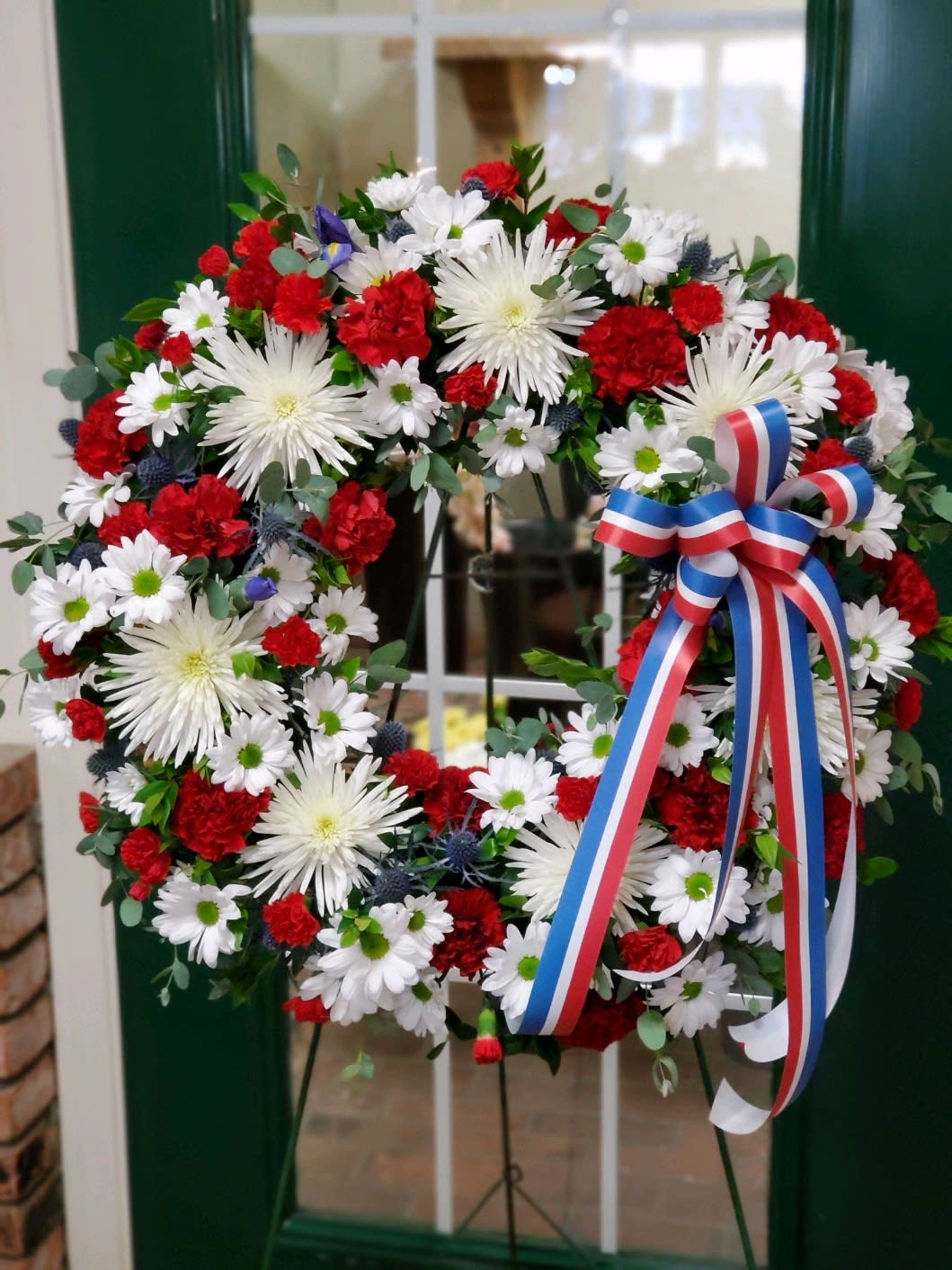 Patriotic Wreath  - Wreath on an easel with red, white and blue flowers to include, Chrysanthemums, carnations, daisies, thistle, greenery with a red, white and blue bow