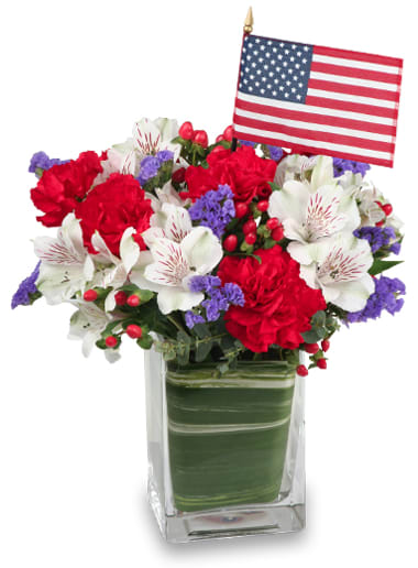 Made in The USA - The Made in The USA bouquet features: Variegated Aspidistra Leaf, Red Grand Slam Carnations, White Virginia Alstroemeria, Blue Statice, Red Hypericum, and a Miniature American Flag artfully arranged in a rectangular glass vase. 