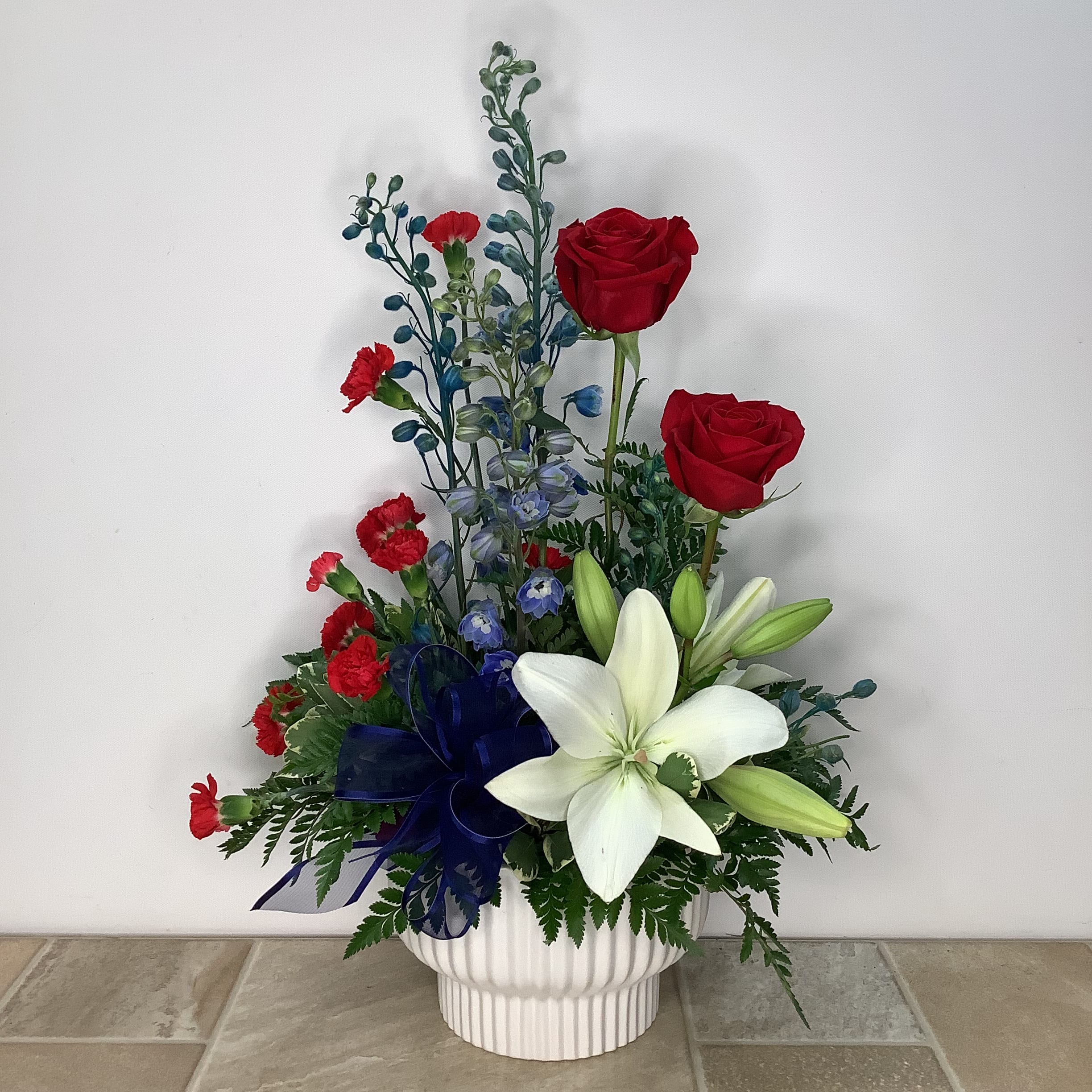 Steadfast - This artistic bouquet of red, white and blue blooms is both patriotic and beautiful. It features red roses and mini carnations, blue delphinium and white lilies all arranged in a white ceramic container. It measures 15” wide x 23” tall.