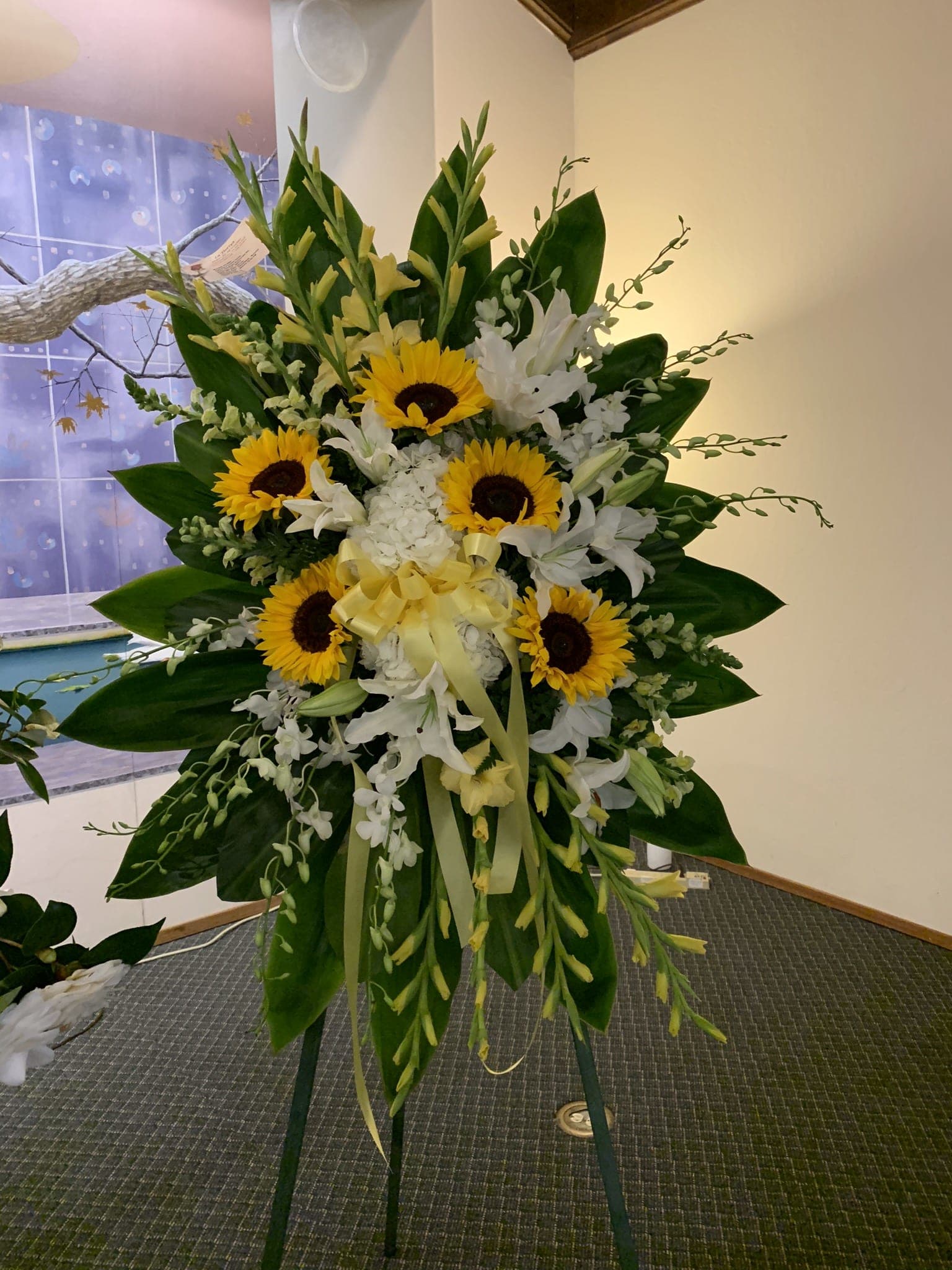 SS#20 Sunny Yellow and White Spray - Send your deepest condolences with these tropical sympathy flowers for the service. The bright yellows and peaceful whites in this standing spray create an impressively balanced display. This beautiful spray includes sunflowers, yellow gladioli, yellow snapdragon, white hydrangeas, dendrobium orchids and oriental lilies and ti leaves.  Orientation: One-Sided  The arrangement is approximately 48&quot; H and 24&quot; W.  Standard (as shown)