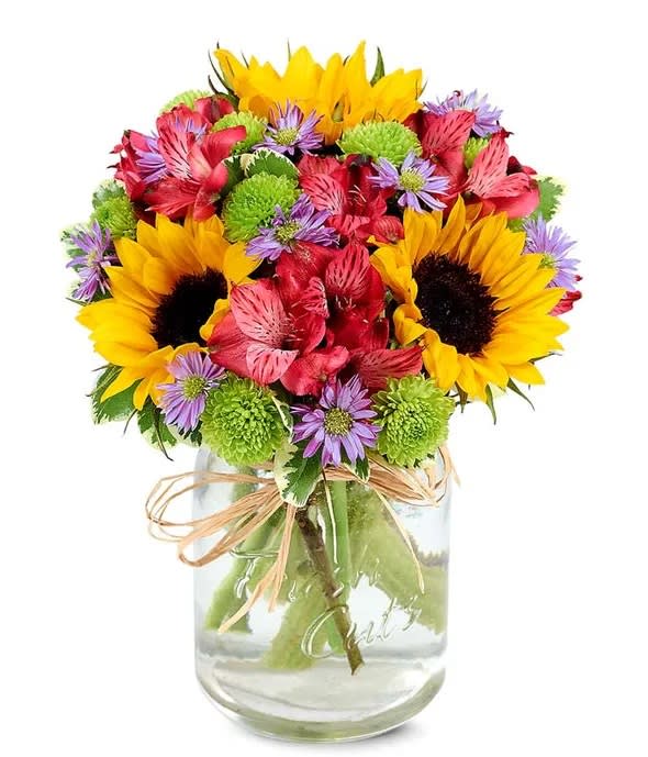 Flower Fields Mason Jar - It's the beauty of a field of flowers from the comfort of home. This bouquet of flowers has a chic farmhouse style while still staying modern. Bright sunflowers surrounded by pink alstroemeria and green poms are displayed in a mason jar wrapped in raffia. The essence of a field of flowers can be brought to anyone's home, and will charm anyone who sees it!