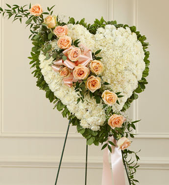 White Heart with Peach Rose Break - This solid heart standing spray, accented with a slash of beautiful peach roses, is a timeless symbol of your love, sympathy and support. White carnations, peach roses, heather, and greenery Solid heart with oasis in mache