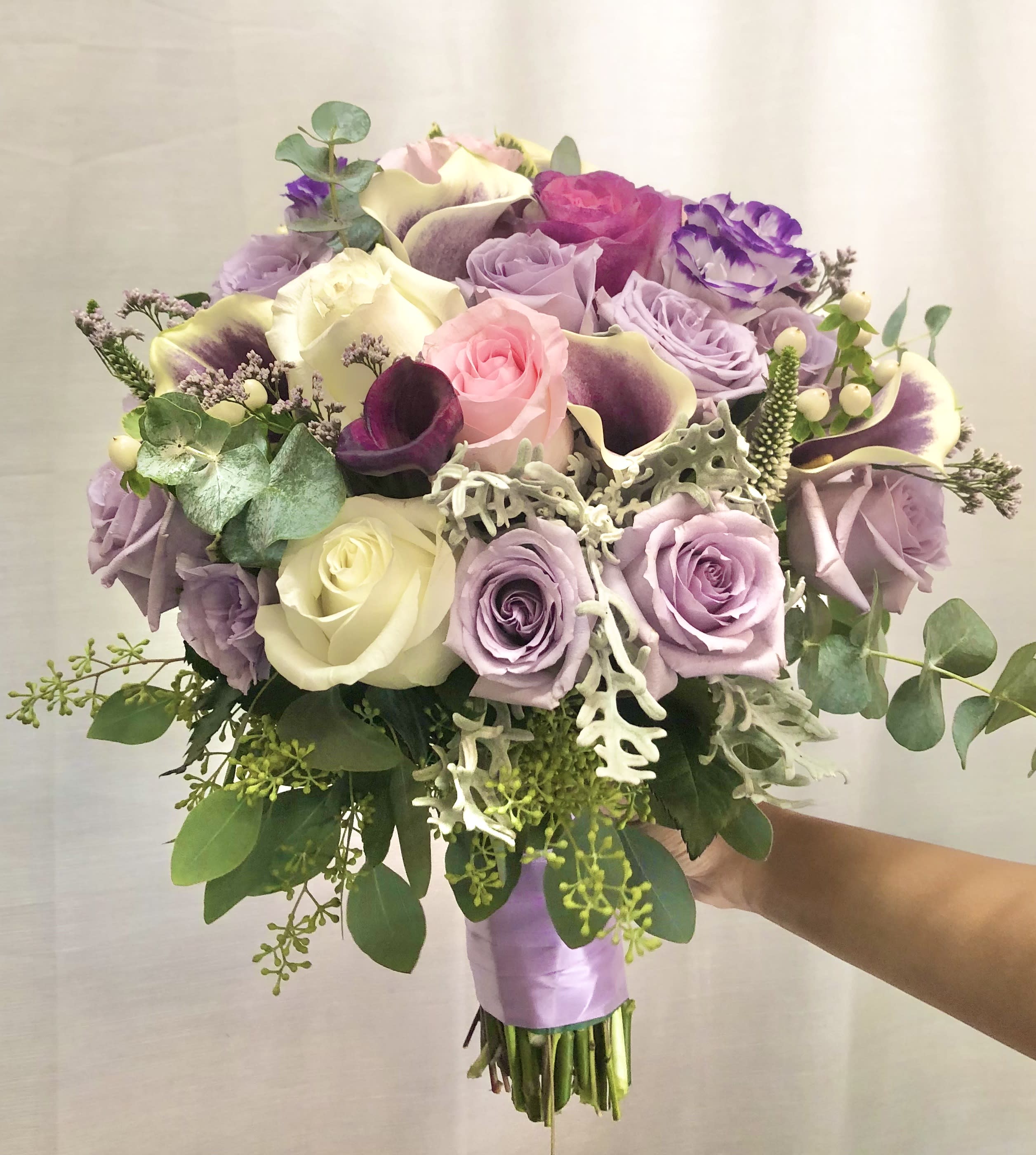 Enchanted  - Beautiful bouquet arrangement with mixed lavender, pink, and white flowers with silver and green accents. Featuring calla lilies, lisianthus, roses, tulips, perennial leaves, and white/green accents. Stems wrapping in bridal bouquet style lavender ribbon. 