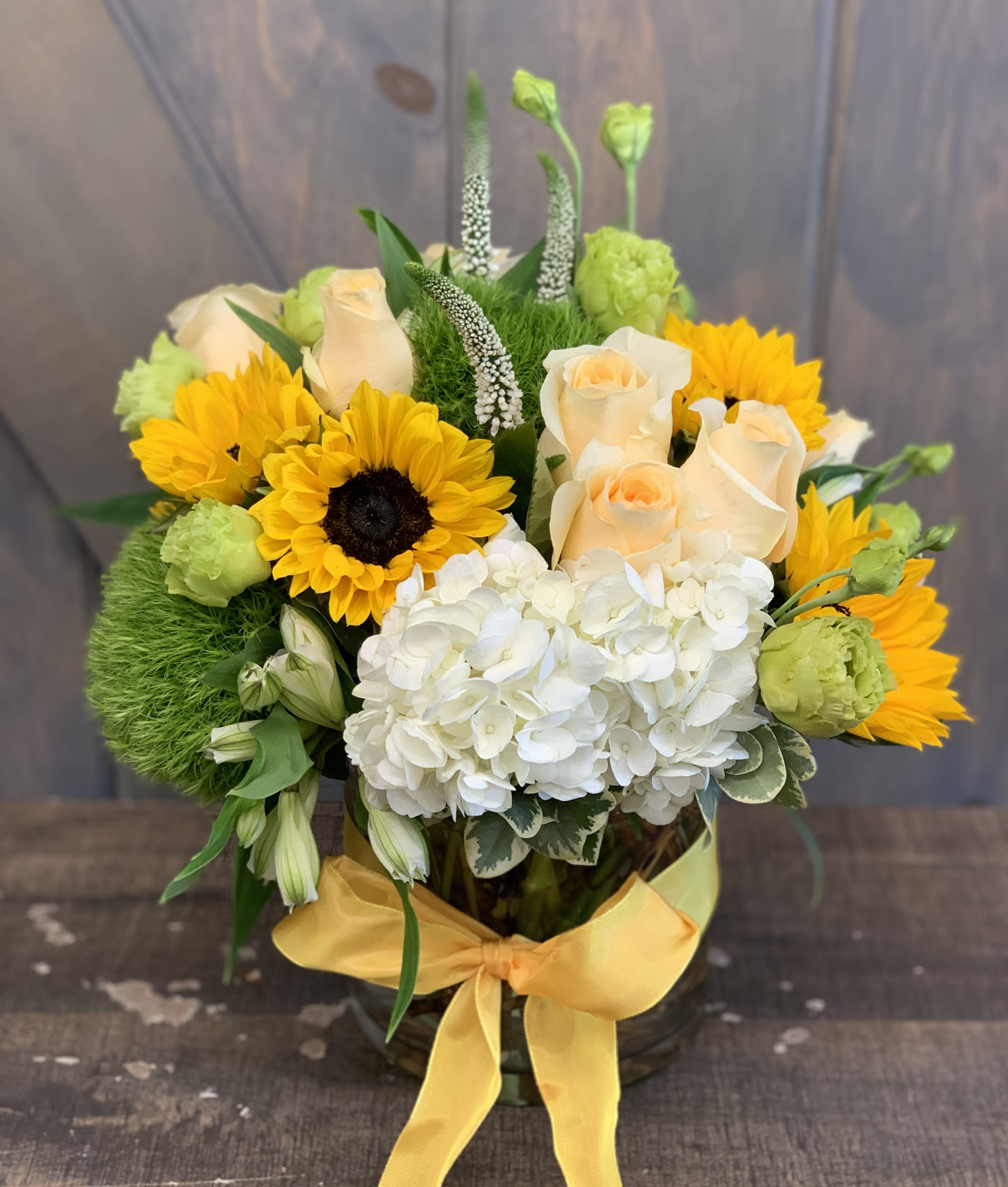 Sunshine Sunshine - This elegant and cheerful arrangement is bond to bring a smile to your recipients face. Included in this design are Hydrangea, Roses, Dianthius, Sunflowers, Veronica, Lisianthius, and Alstromeria designed in a 6x6in clear glass vase
