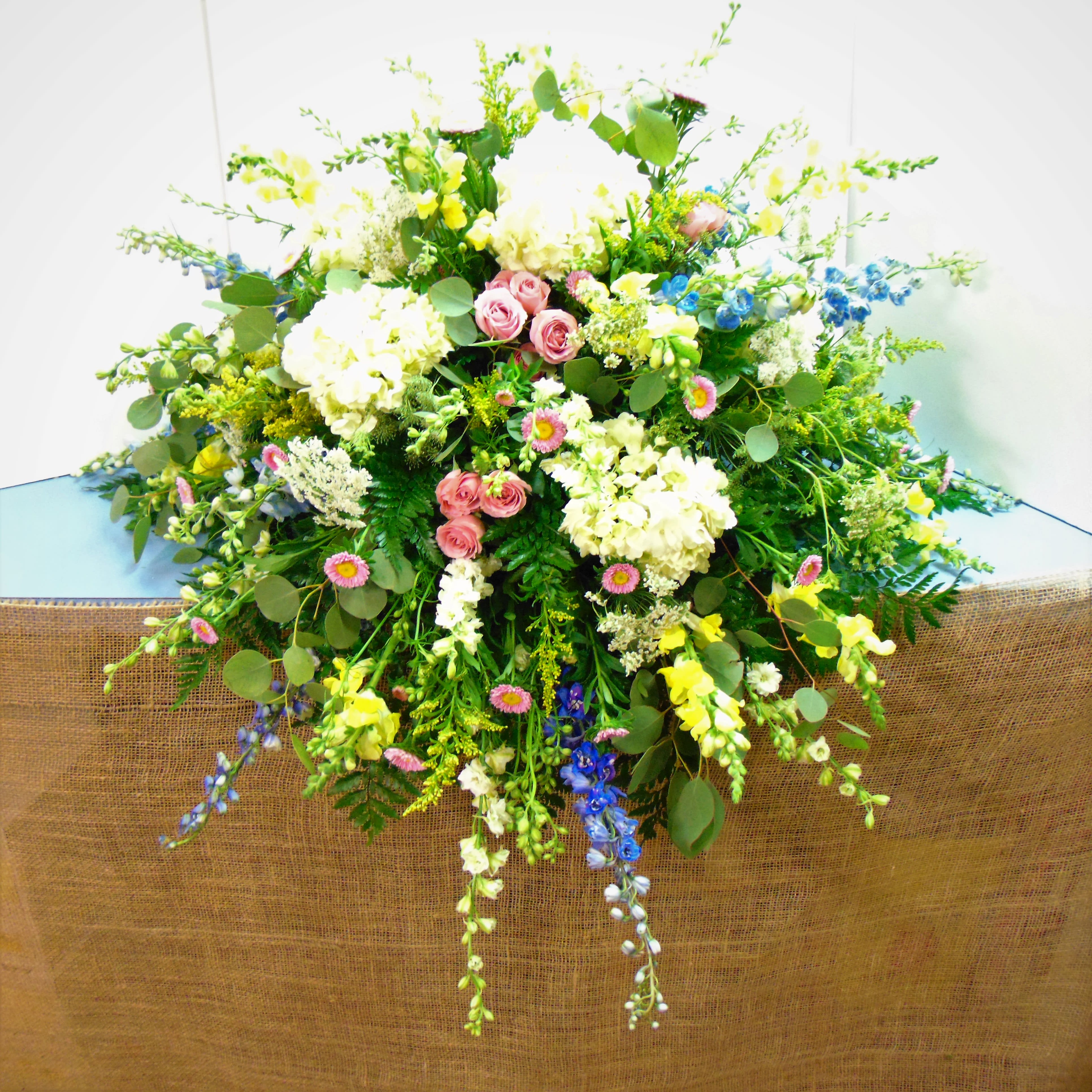 Pastel Perfection Casket Spray - Soft, restful blend of light blue hydrangea, yellow snapdragons, blue delphinium, pink spray roses and garden style accents with greenery. Standard option is pictured. Deluxe option adds more pink spray roses, white and yellow daisies.  The premium option adds 12 stems of yellow and pink spray roses, more blue delphinium and queen anne's lace.