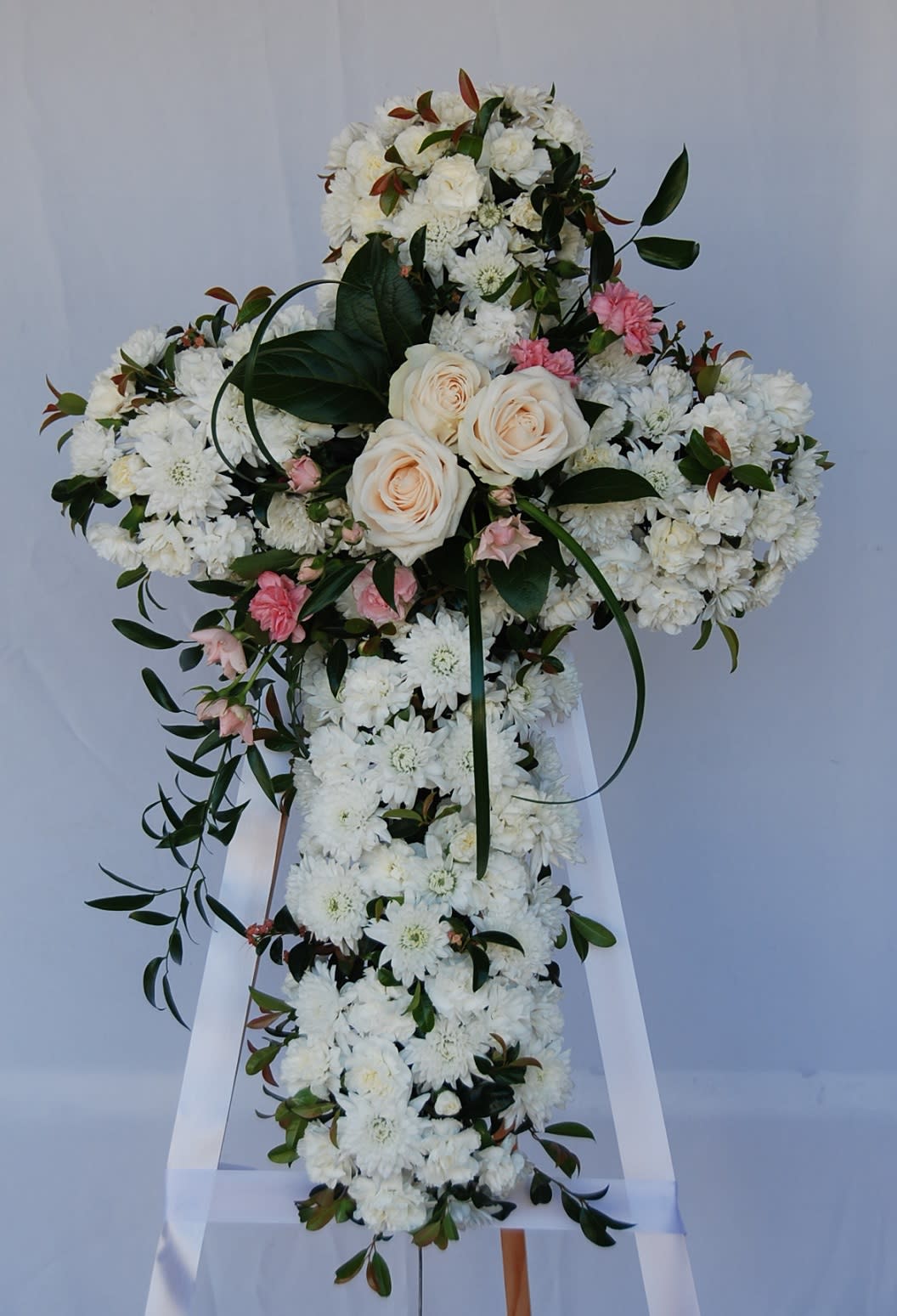 White Cross - Beautiful flowers such as crème roses, white spray roses, carnation, leptospermum, cushion and button spray chrysanthemums on an easel-mounted standing cross. Orientation: One-Sided