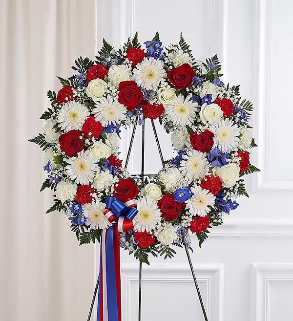 Serene Blessings™ Standing Wreath- Red, White &amp; Blue - They served their country with honor and pride, so it’s only fitting to honor them with a beautiful symbol of eternal life. Our patriotic standing wreath arrangement is meticulously crafted by our expert florists to honor a brave veteran who has passed away. Filled with lush blooms in red, white and blue, it’s a fitting final tribute for the funeral services.