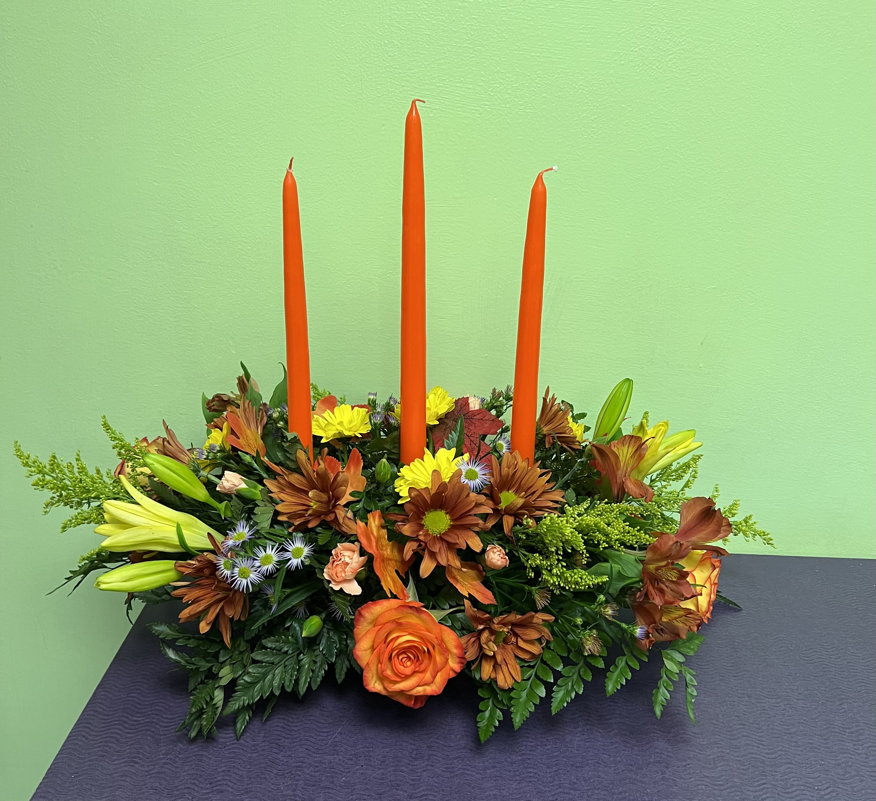 Magnificent Thanksgiving Centerpiece - This 3 candled centerpiece will fill your Thanksgiving table will much needed fall blooms. This large centerpiece includes Lilies, Roses, Chrysanthemums, Aster and some fall leaves. 