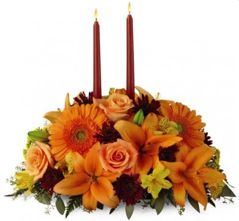 B44112 - Bright Autumn This Centerpiece spins the magic of the Fall season with each sun-kissed petal to set your gathering space aglow with blooming beauty. Two taper candles are surrounded by a gorgeous arrangement consisting of orange Asiatic Lilies, gerbera daisies, and roses with golden Peruvian Lilies, burgundy cushion poms and assorted greens to create the perfect addition to your Autumn celebration.