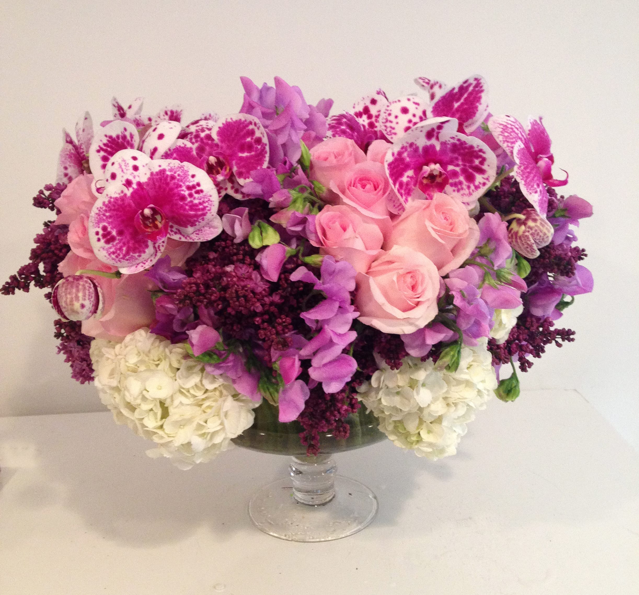 Wisteria Lane - A lush array of roses, sweet pea, phalaenopsis orchids, and lilacs in shades of pale pinks, deep purples, and lavender in a decorative pedestal glass vase. This is a fragrant and very boutique arrangement that is full of seasonal flowers, please allow at least one day for delivery.  *vase subject to availability