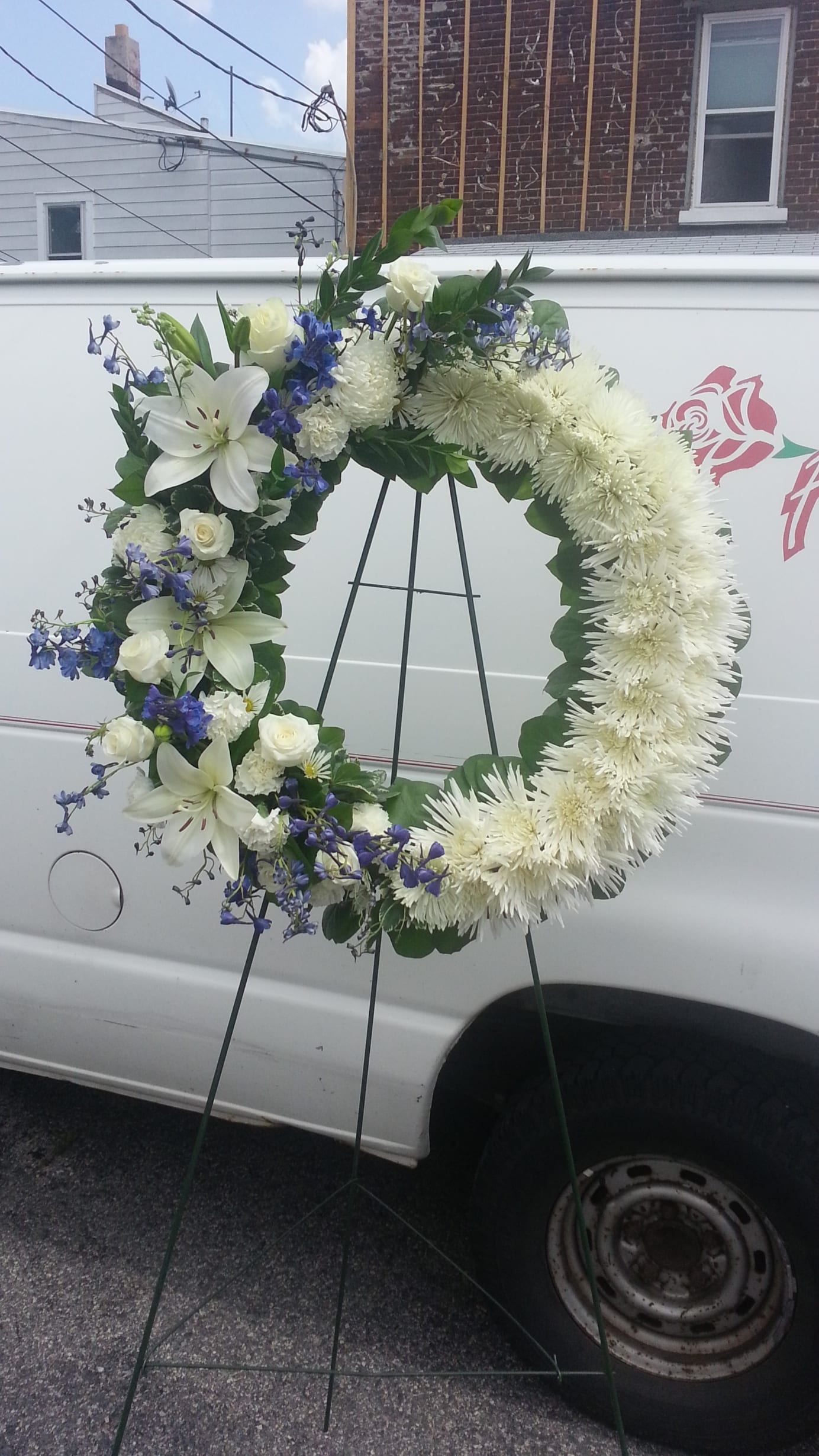 A Touch of Blue - Display your thoughts with this wreath covered in large white mums, with some carnations, roses, lilies, and added touch of some blue delphinium