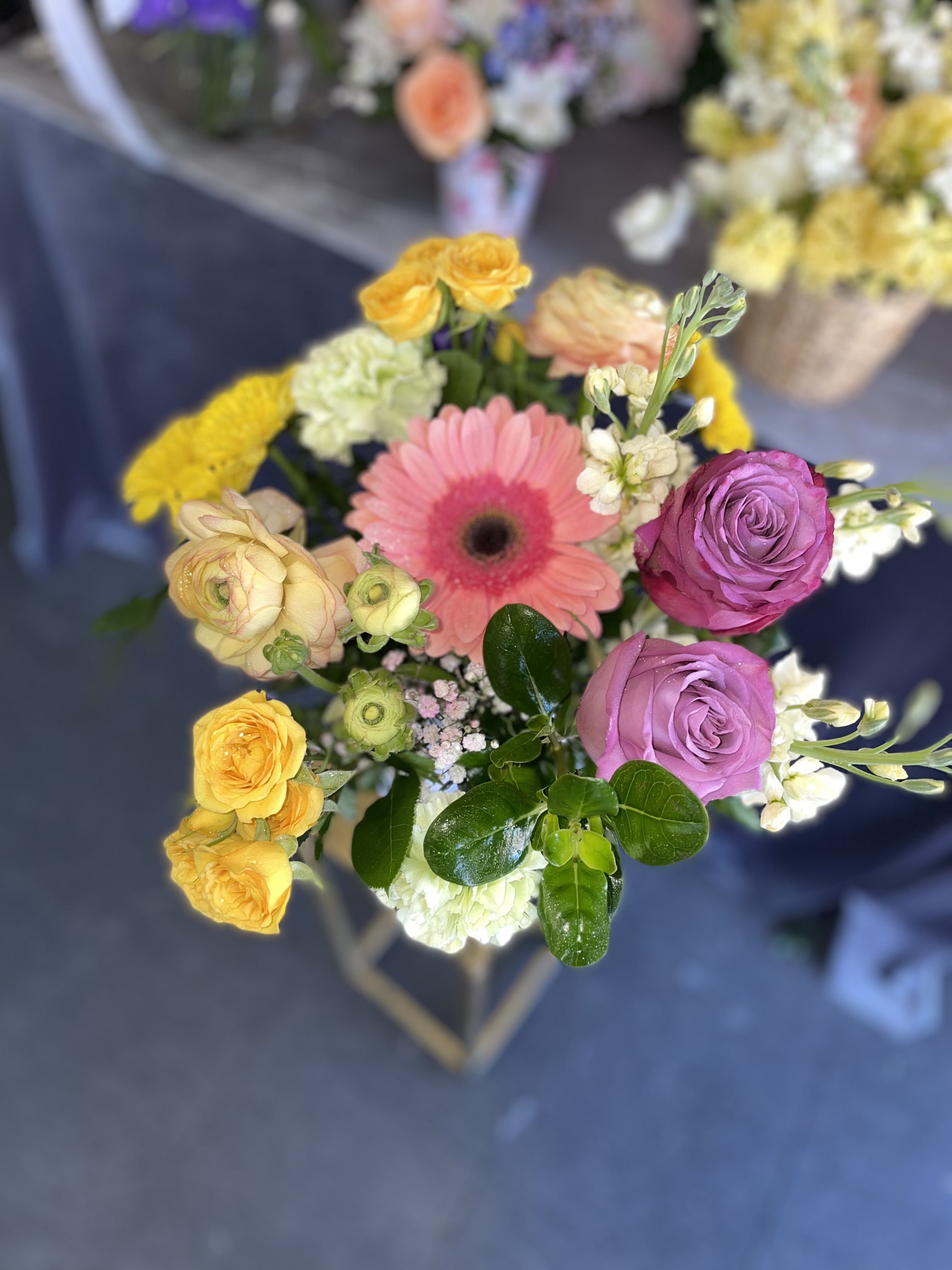 Blooming - This beautiful arrangement has some nice colors and fresh flowers, this arrangement has yellow spray roses, lavenders roses, pink gerbera, yellow pompos, peach ranunculus, green carnations, and fresh greens.