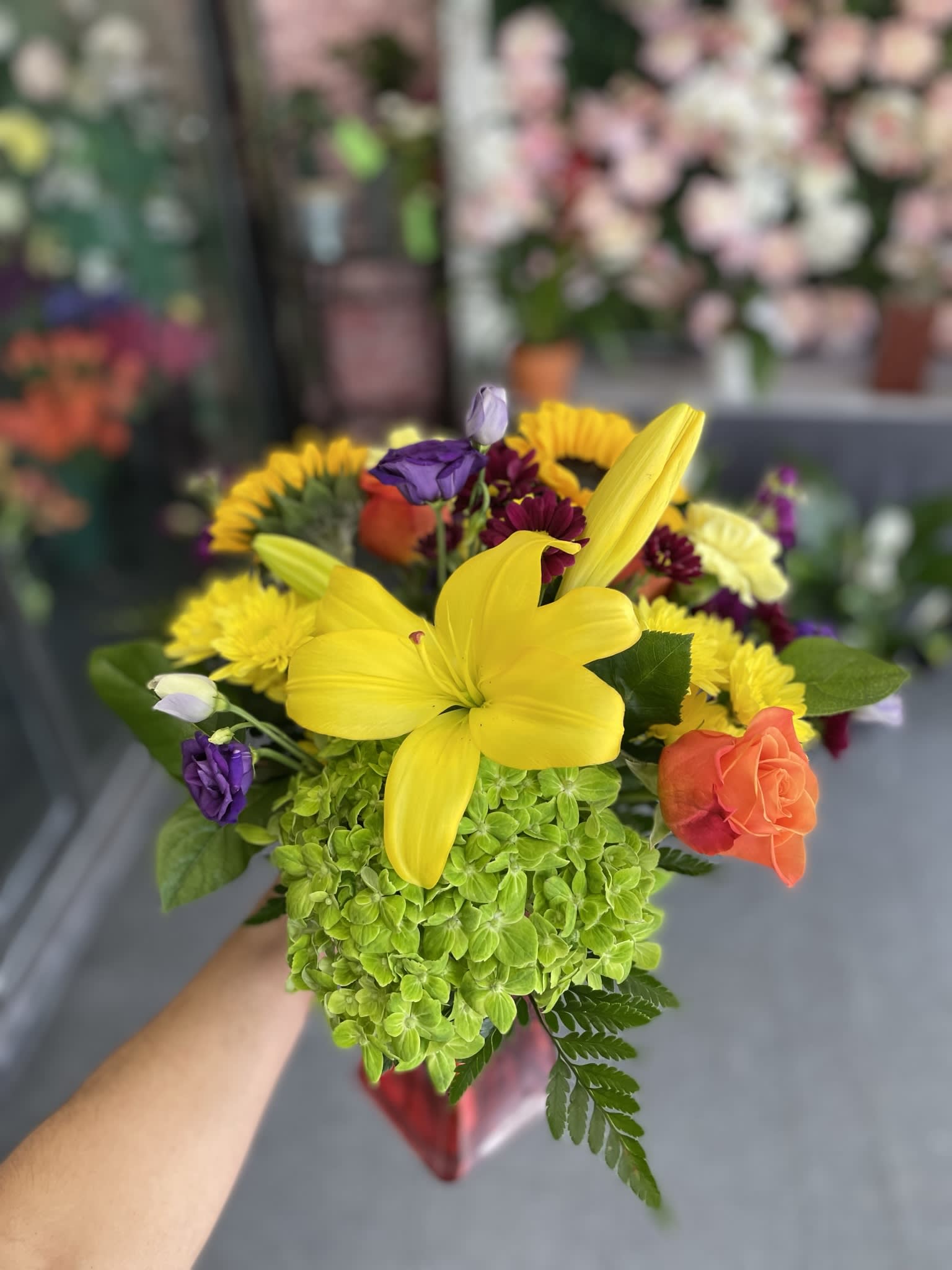 Bella Fluer - This beautiful arrangement has green hydrangea, yellow lily’s, orange roses, sunflowers, purple lusy, stock, yellow carnations, and a mix of pompos.