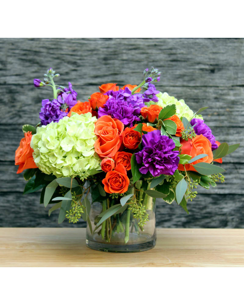 Vibrant Gratitude - This bright and vibrant arrangement will liven up any room. Orange roses &amp; mini roses, purple carnations &amp; stock and green hydrangea with assorted greens in a clear glass cylinder vase.