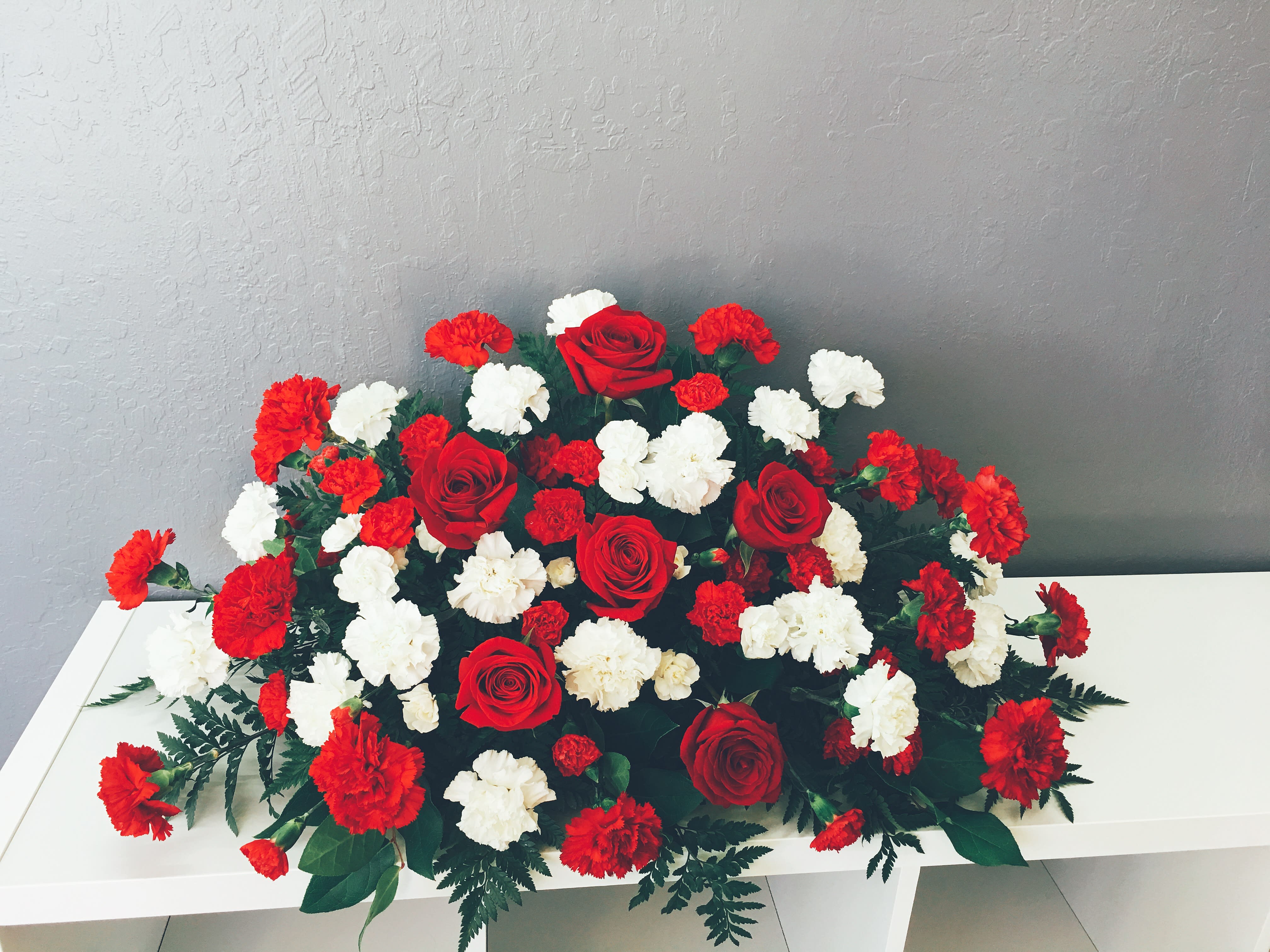 CS#11 Red &amp; White Carnation Casket Spray - Red carnations to symbolize affection and white carnations to symbolize purity, a simple and powerful statement to pay tribute to a loved one.  The standard (half-open casket) arrangement is approximately 38” W x 16” H. The deluxe (closed casket) arrangement is approximately 46” W x 16” H.