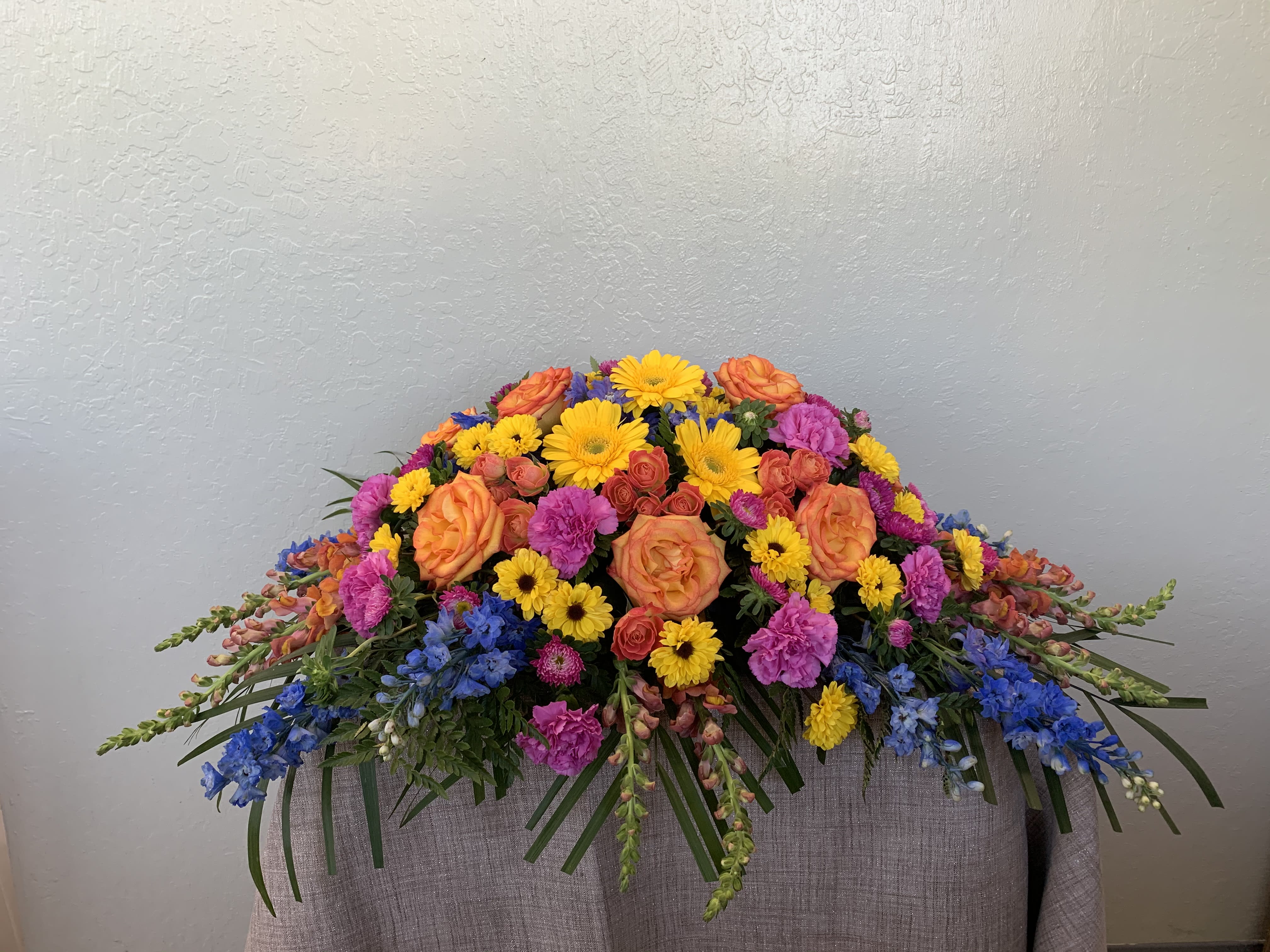 CS#15 Celebration of Life Casket Spray - A colorful assortment of flowers with a spring appearance featuring gerbera daisies, roses, carnations, snapdragons, spray roses, delphinium, filling flowers, and greens.  Standard as shown (half-open casket) approximately 36” W x 16” H Deluxe (closed casket) approximately 44” W x 16” H