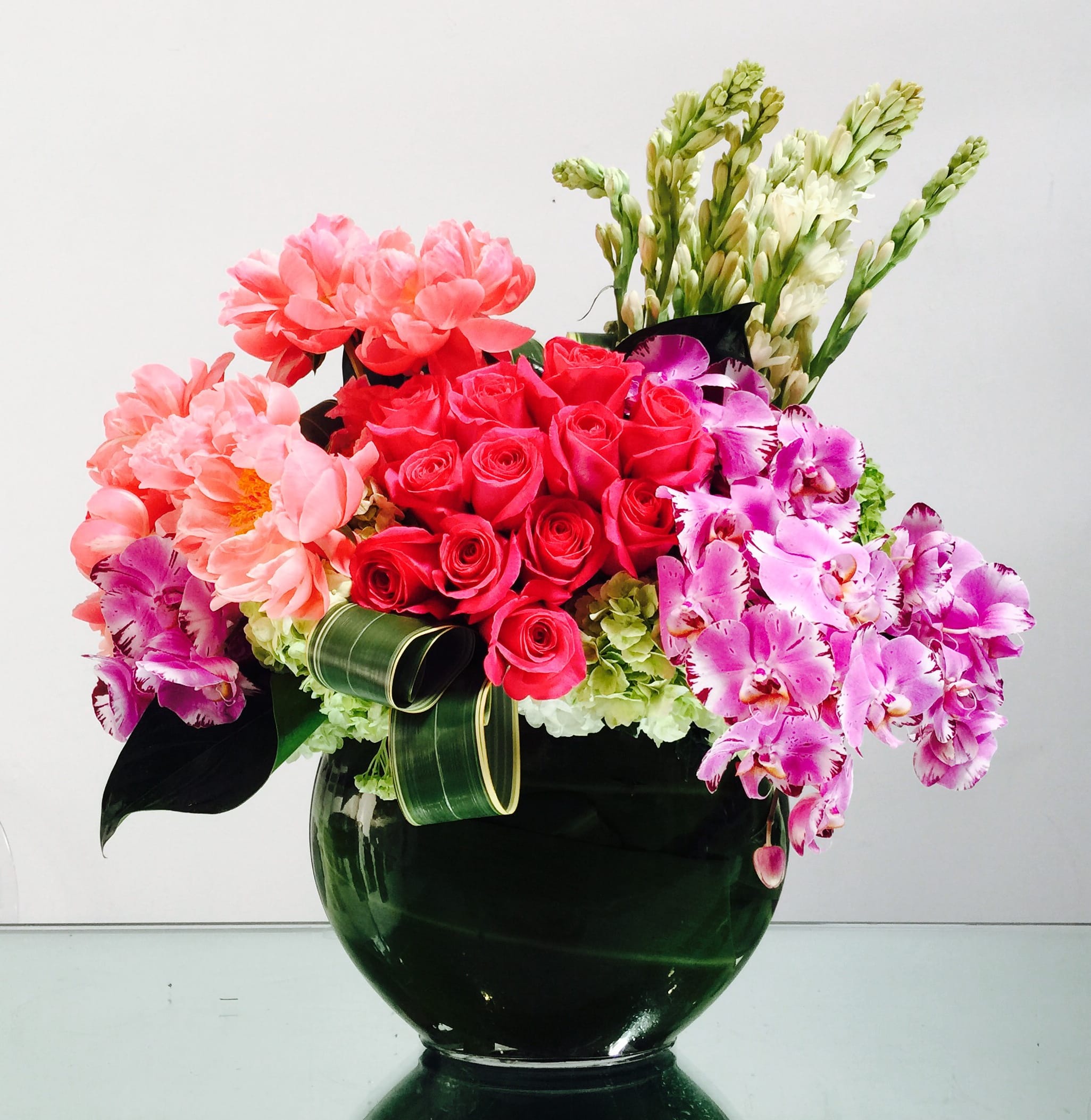 Peony Pink Peony Pink - Coral peonies stand out against fuschia roses, magenta phalaenopsis orchids, an accent of tuberose, and greens. This fragrant arrangement in a 10" leaf-lined bubble bowl will be an olfactory delight!