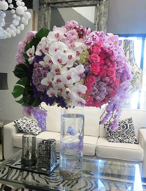 Cassanova - A Grand Centerpiece Arrangement. An opulent mixture of white and lavender orchids, hot pink and purple orchids, white hydrangeas, monstera leaves atop a Glass Cylinder vase with Submerged Orchids. 