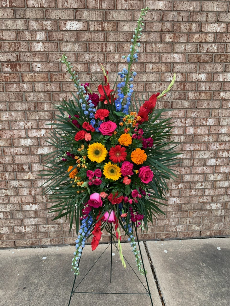Celebration of Life standing spray - A standing spray that shows off every selection of the spectrum, with vibrant reds, oranges, yellows, blues, purples, and pinks with foliage accents atop an easel. A colorful combo of gladiolus, gerbera daisies, delphinium, roses, stock, and carnations, this mix is the ideal way to pay thoughtful tribute to a loved one who brings bright memories of cheer and happiness to mind.