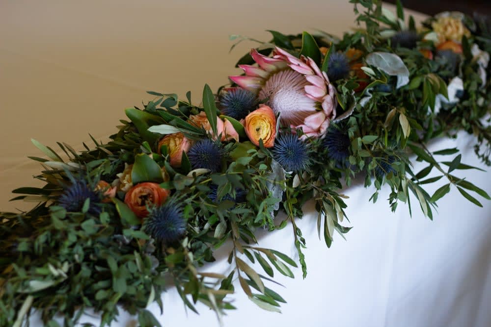 Judy - This garland is both striking and elegant. Using the boldness of protea and the sturdiness of thistle as well as other seasonal flowers and greens, this earthy flower arrangement is great on banners, tables, balconies...etc. 