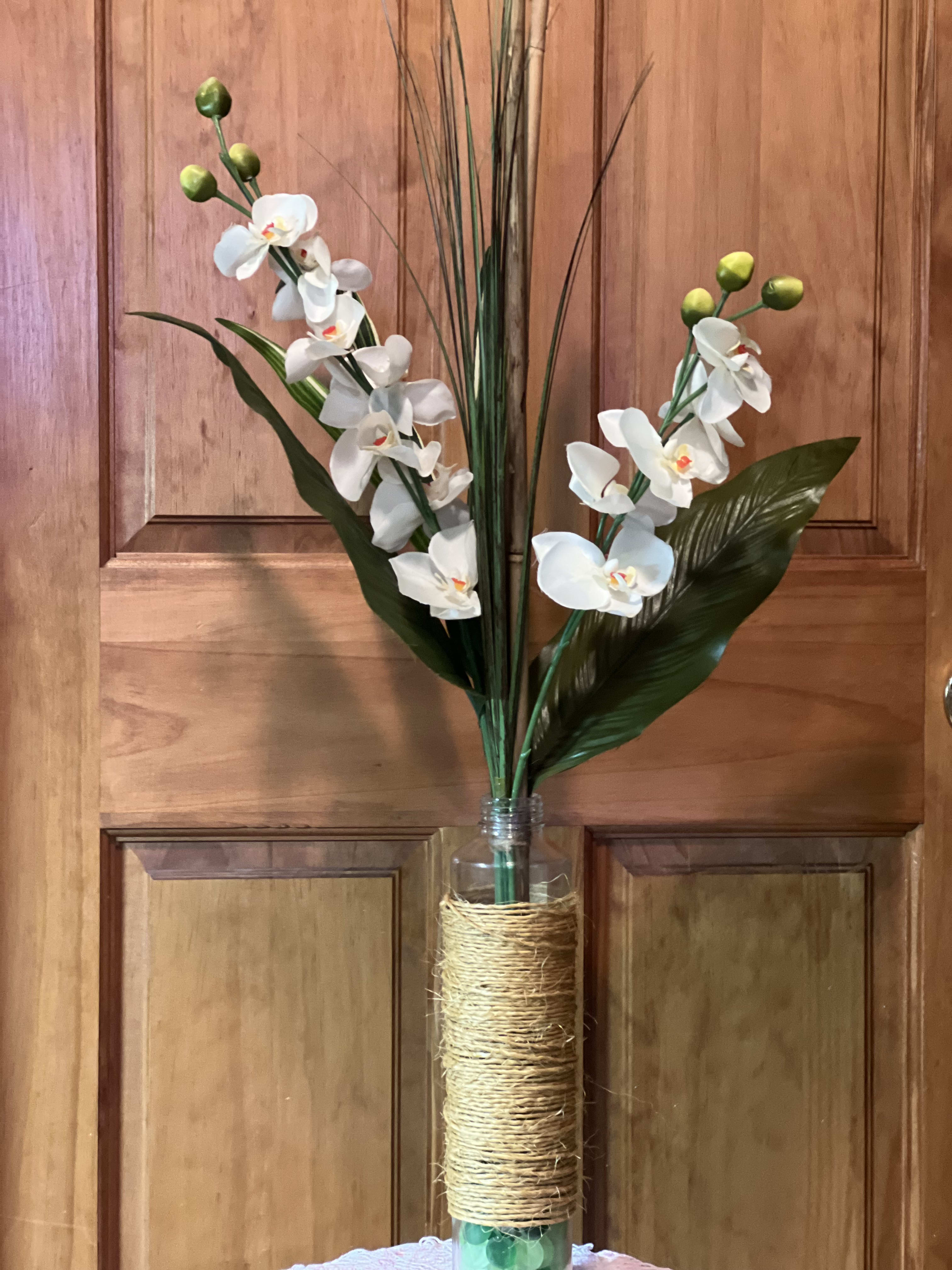 Tropical Kiss - Our One-of-a-Kind Tropical Kiss is a long lasting silk arrangement tied together with twine around the base and the stems to keep everything together. Permanent silk orchids, bamboo, assorted ti leaves and bear grass with glass fillers inside the clear vase. Great for an entryway, hall table, or displayed in a corner. Overall height: 30&quot; tall 