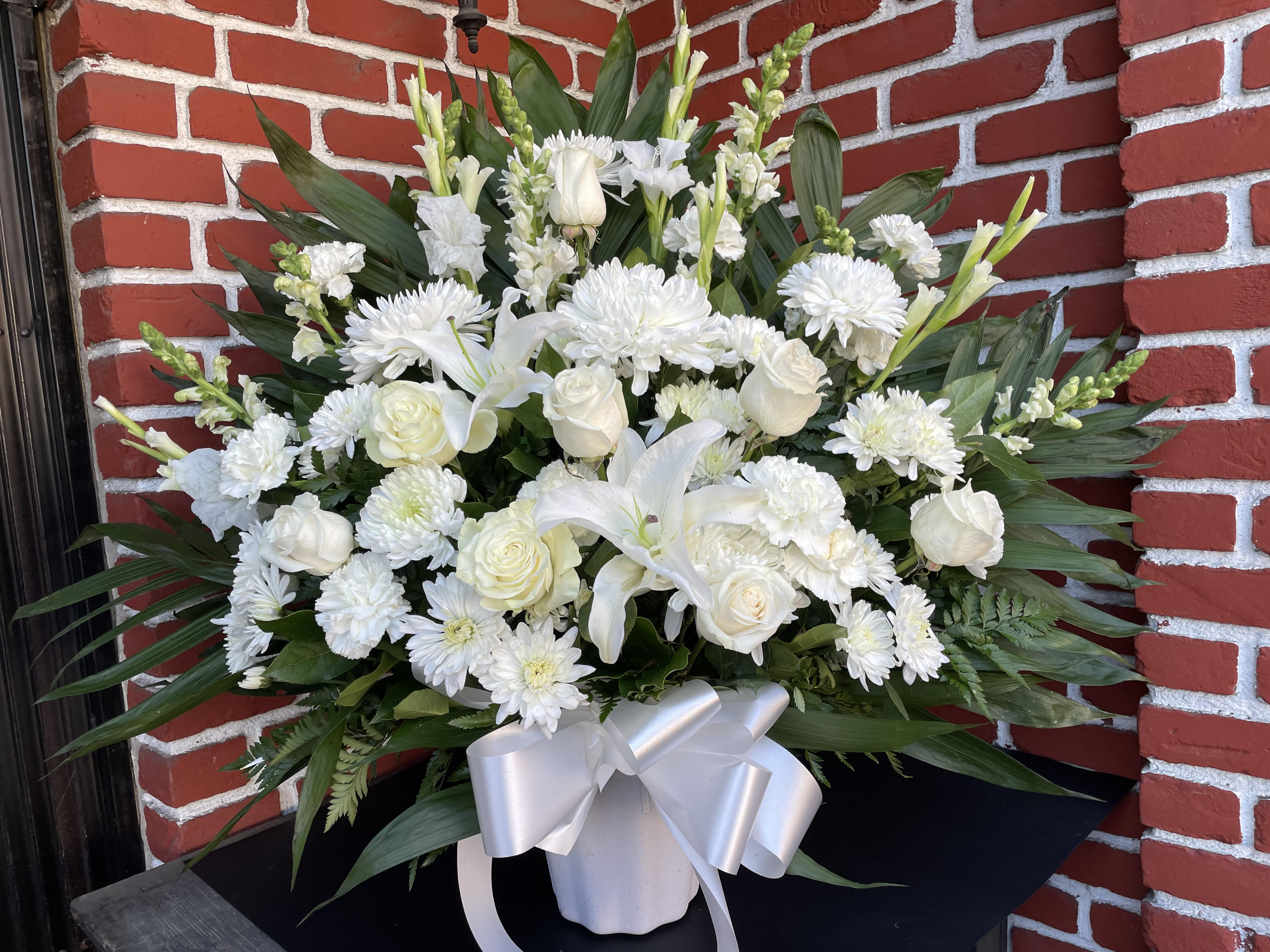 Rose and Lily White funeral basket  - An all-white funeral basket featuring  lilies, roses, dragon snaps, and more framed by a backdrop of large leaves. Suitable for any type of funeral ceremony