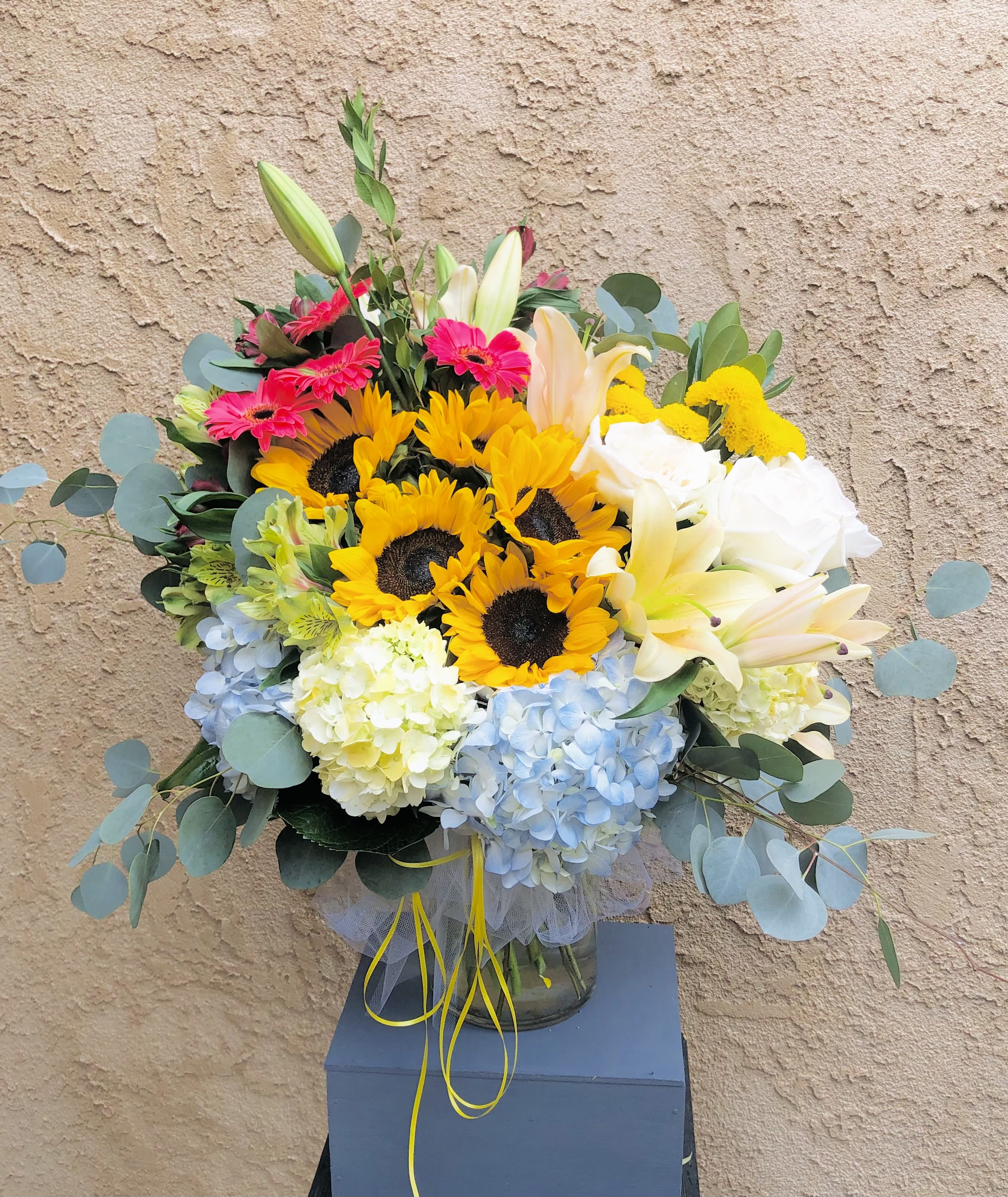 Everyday Warmth - Beautiful yellow, blue, white, and pink vase arrangement featuring sunflowers, daises, hydrangeas, roses, and lilies with green leaf accents.