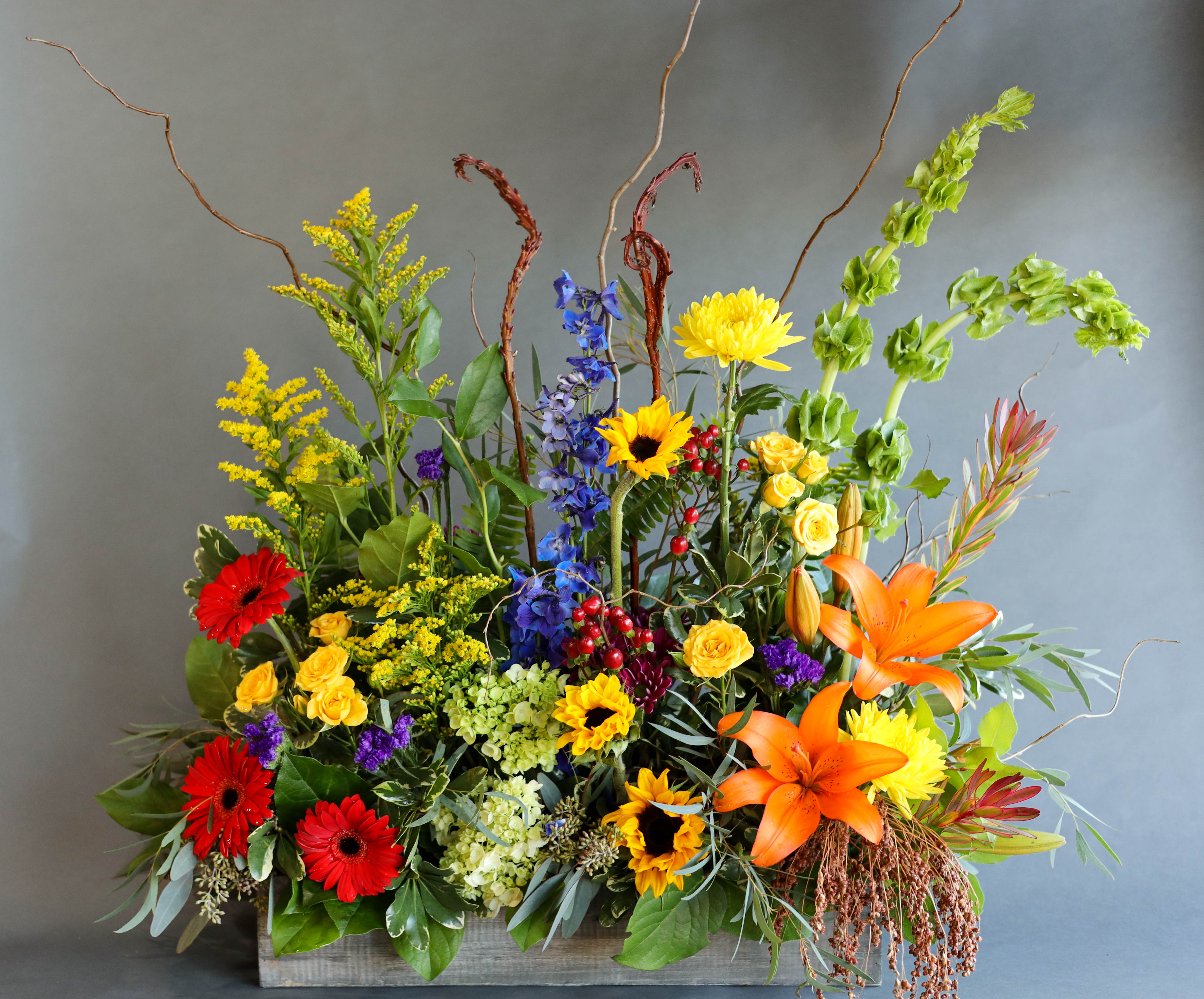 Warm Thoughts of Sympathy - A beautiful lush arrangements standing tall with a plethora of our flower varieties like Sunflowers, Lilies, Bells of Ireland, and Gerbera Daisies, to name a few.  