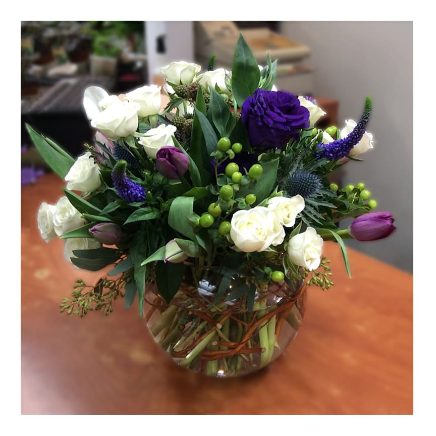 Gracious - Gracious is an elegant two-tone centerpiece. White spray roses, deep purple lisianthus, veronicas, and tulips (seasonal) are accentuated with green hypericum berries, thistle and seeded eucalyptus. Flowers arranged in a glass fishbowl lined with curly willow.  