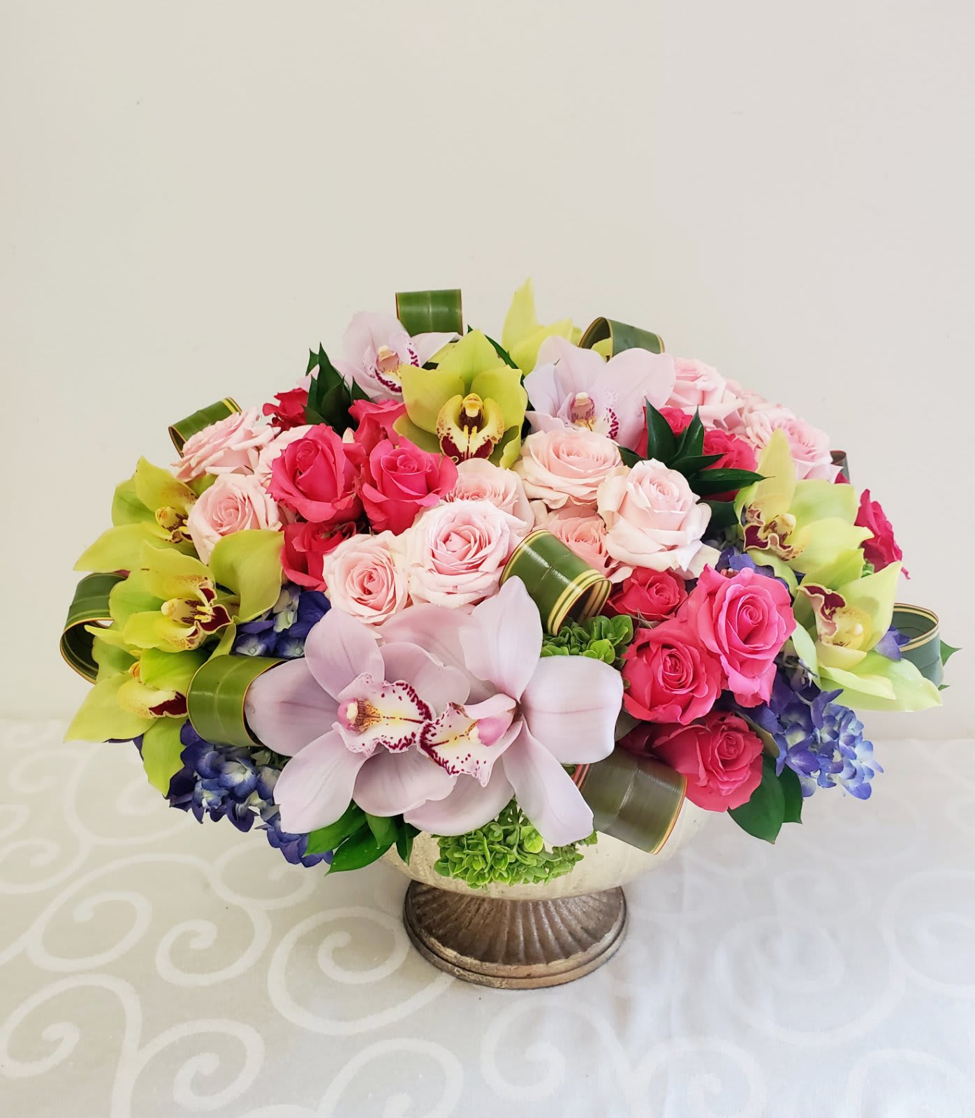 Exotic You | 15"H by 13"W Exotic You | 15"H by 13"W - This collaboration of colors and nature creates a magnificent floral masterpiece in a mixture of colorful Cymbidium orchids and dainty spray roses blended in a bed of purple and green hydrengea finished and accented with beautiful loops of rich green leaves makes a rare and exceptionally stunning piece.